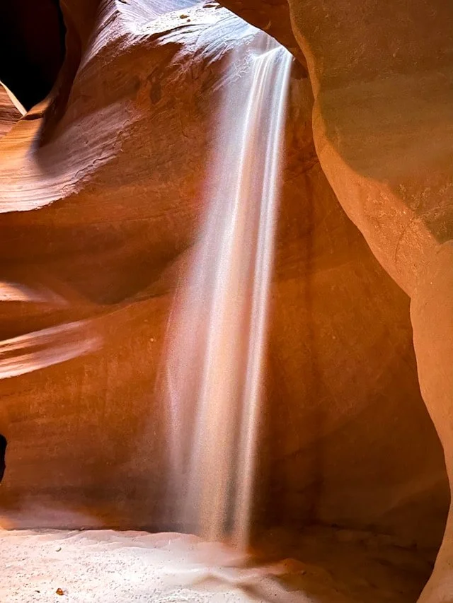 A narrow slot canyon with smooth, reddish-brown sandstone walls and a thin stream of light shining down on a waterfall in the center.