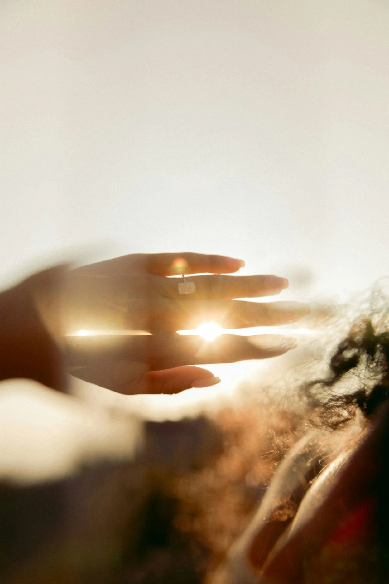 A hand with painted nails and a ring, holding up three fingers against the setting or rising sun, with the sun's light shining through the fingers, and some curly hair visible in the foreground.
