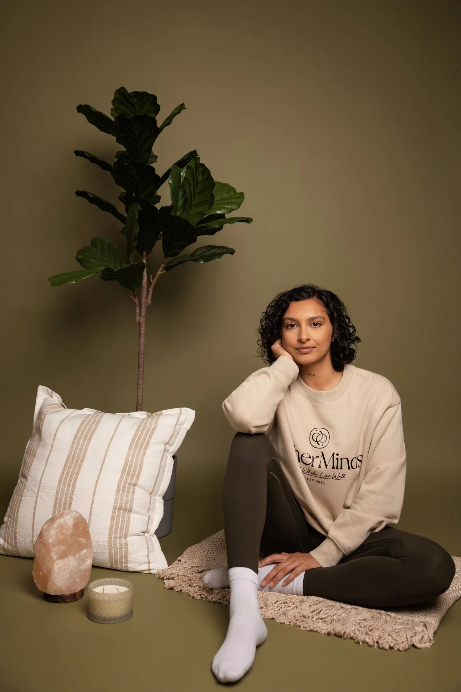 A woman with curly dark hair, sitting on a beige shaggy rug against a green wall, wearing a beige sweatshirt with the logo "Her MInds" and black pants, resting her head on her hand. To her left, a striped pillow, Himalayan salt lamp, candle, and beige blanket are on the floor. Behind her, there is a potted fiddle leaf fig plant.