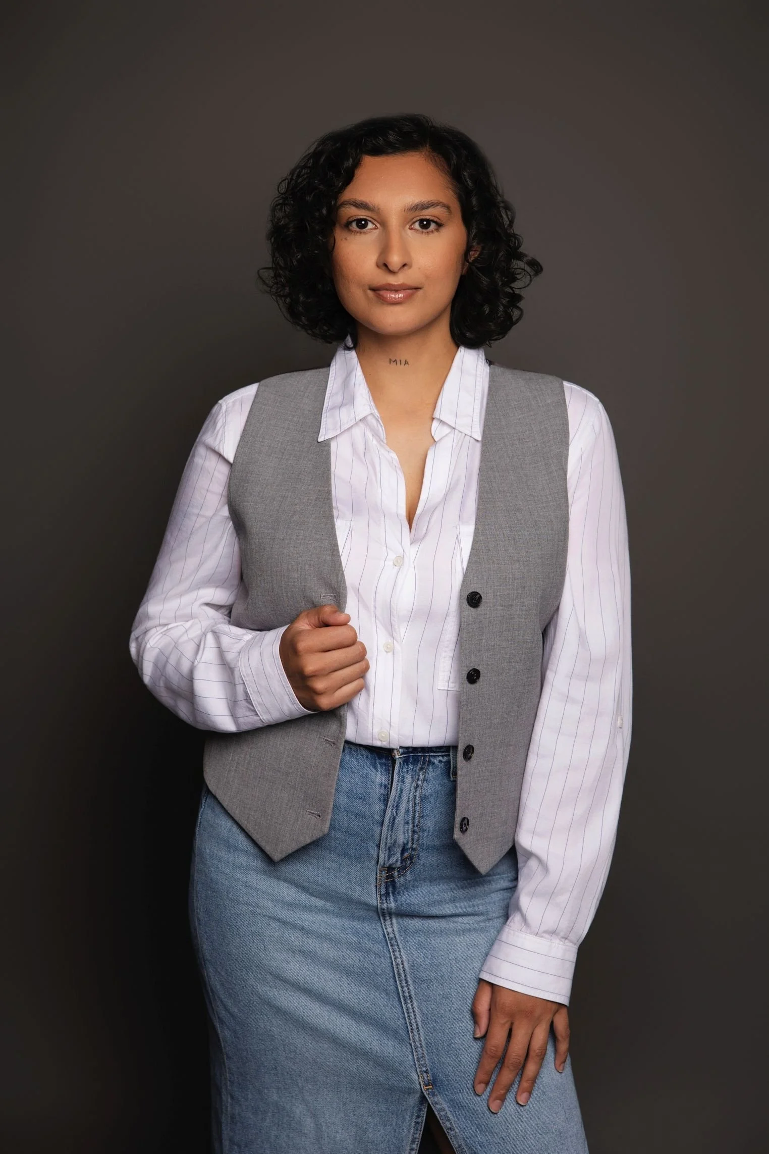 A woman with dark, curly hair standing against a plain dark background, wearing a white shirt with light purple stripes, a gray vest, and blue jeans, holding her vest with one hand.