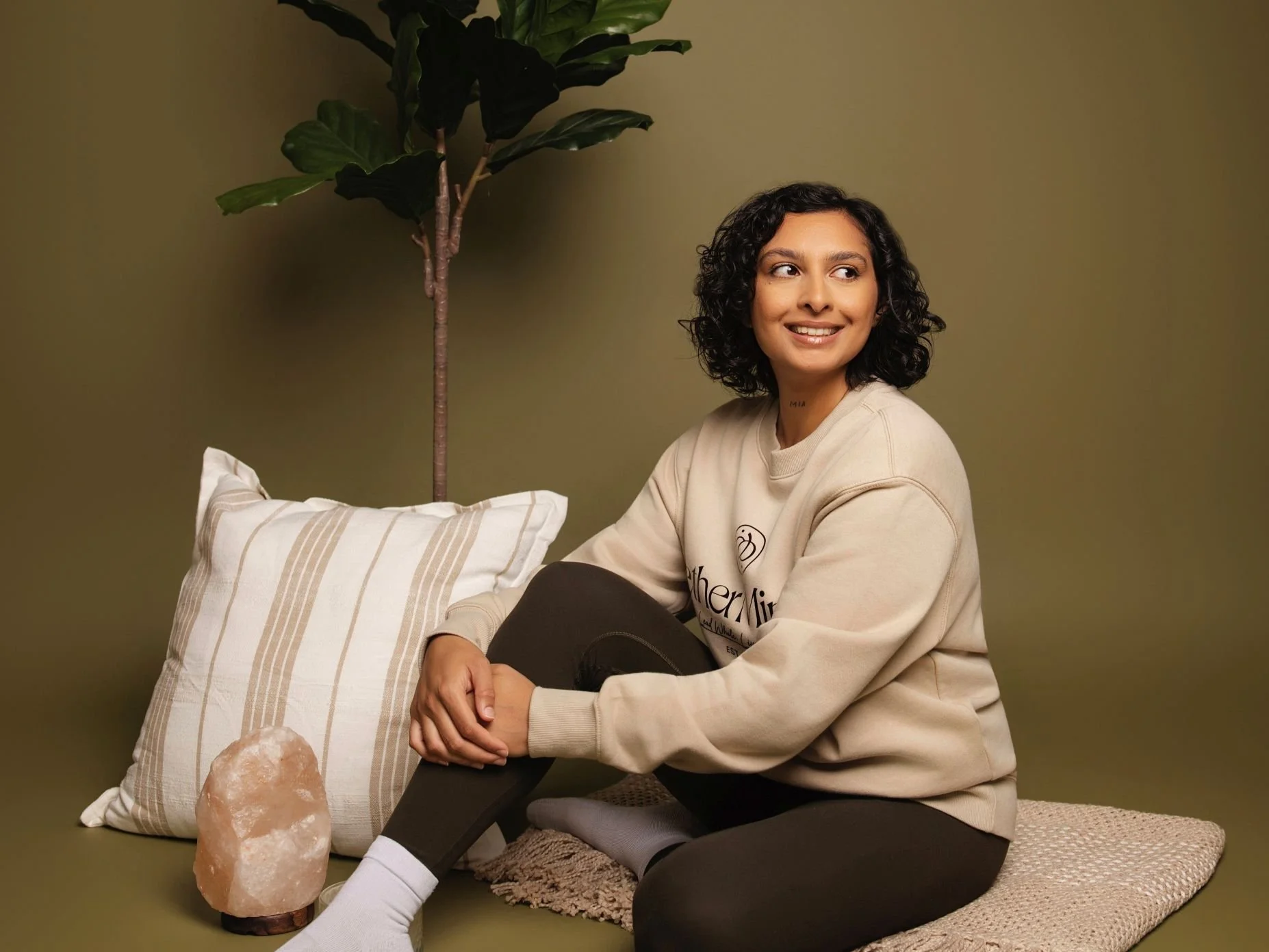 A woman with curly dark hair and a tan sweatshirt, sitting on a woven rug with her legs crossed, smiling and looking to her right. She is in a room with a muted green wall, near a large potted plant with broad green leaves. There is a striped pillow and a large pink salt lamp on the floor.