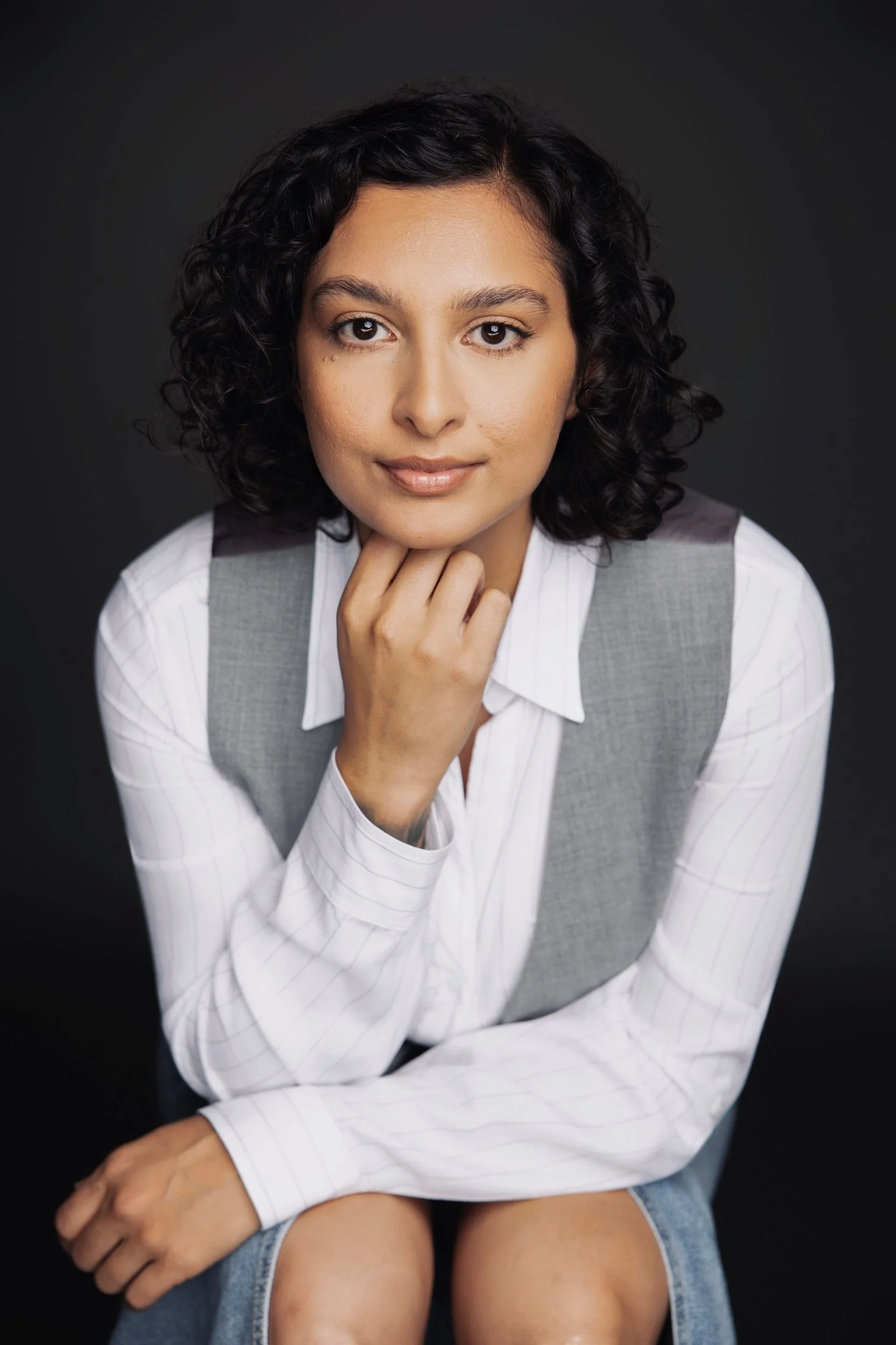 A woman with curly black hair wearing a white shirt with a grey vest, sitting with one hand resting on her chin, against a dark background.