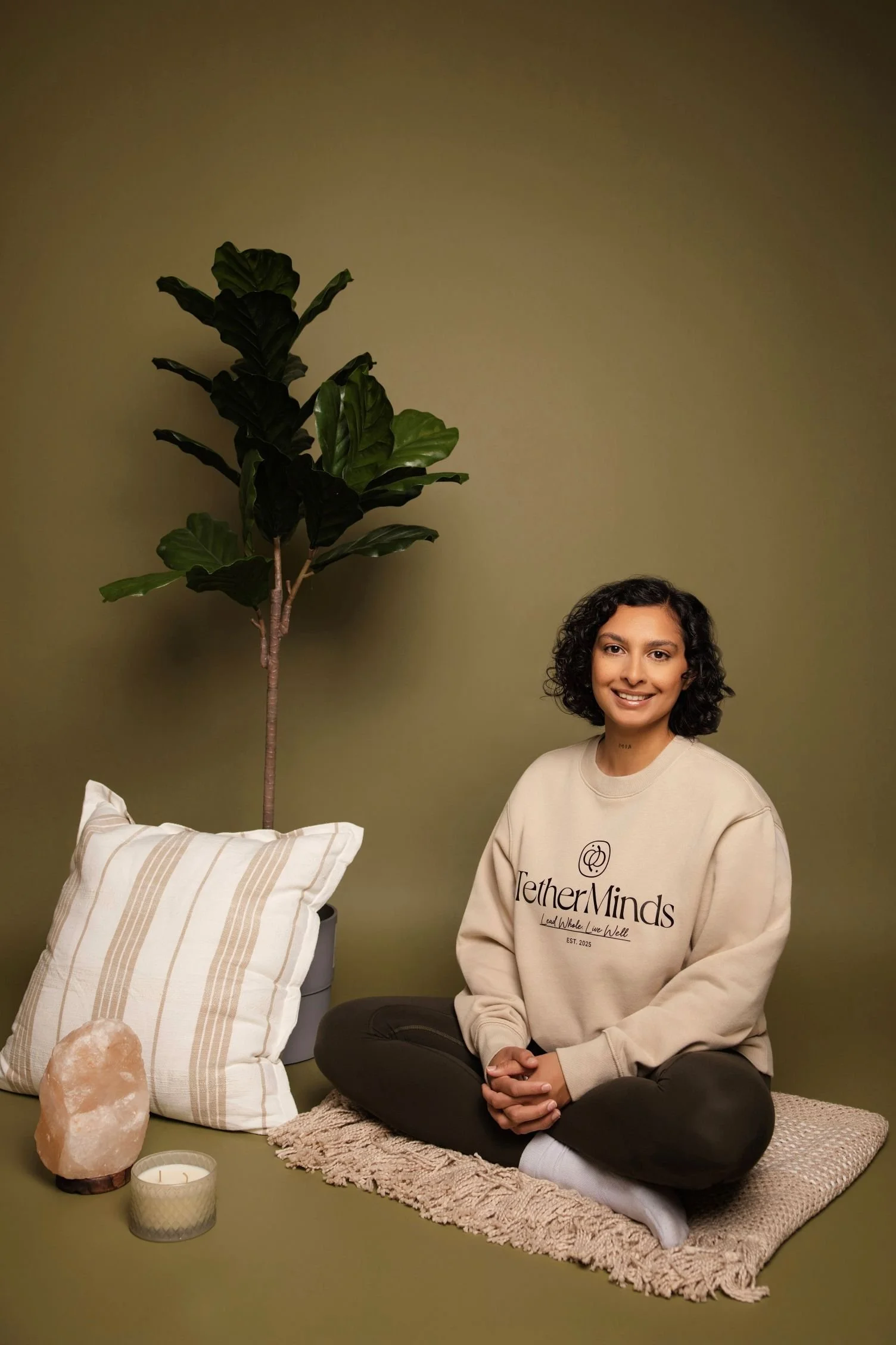 A woman with curly dark hair sitting cross-legged on a knitted beige rug in front of a green wall, wearing a beige sweatshirt with the logo 'TetherMinds' and text. Next to her is a small round pillow with beige stripes, a salt lamp, a white candle in a decorative holder, and a green potted plant with large leaves.