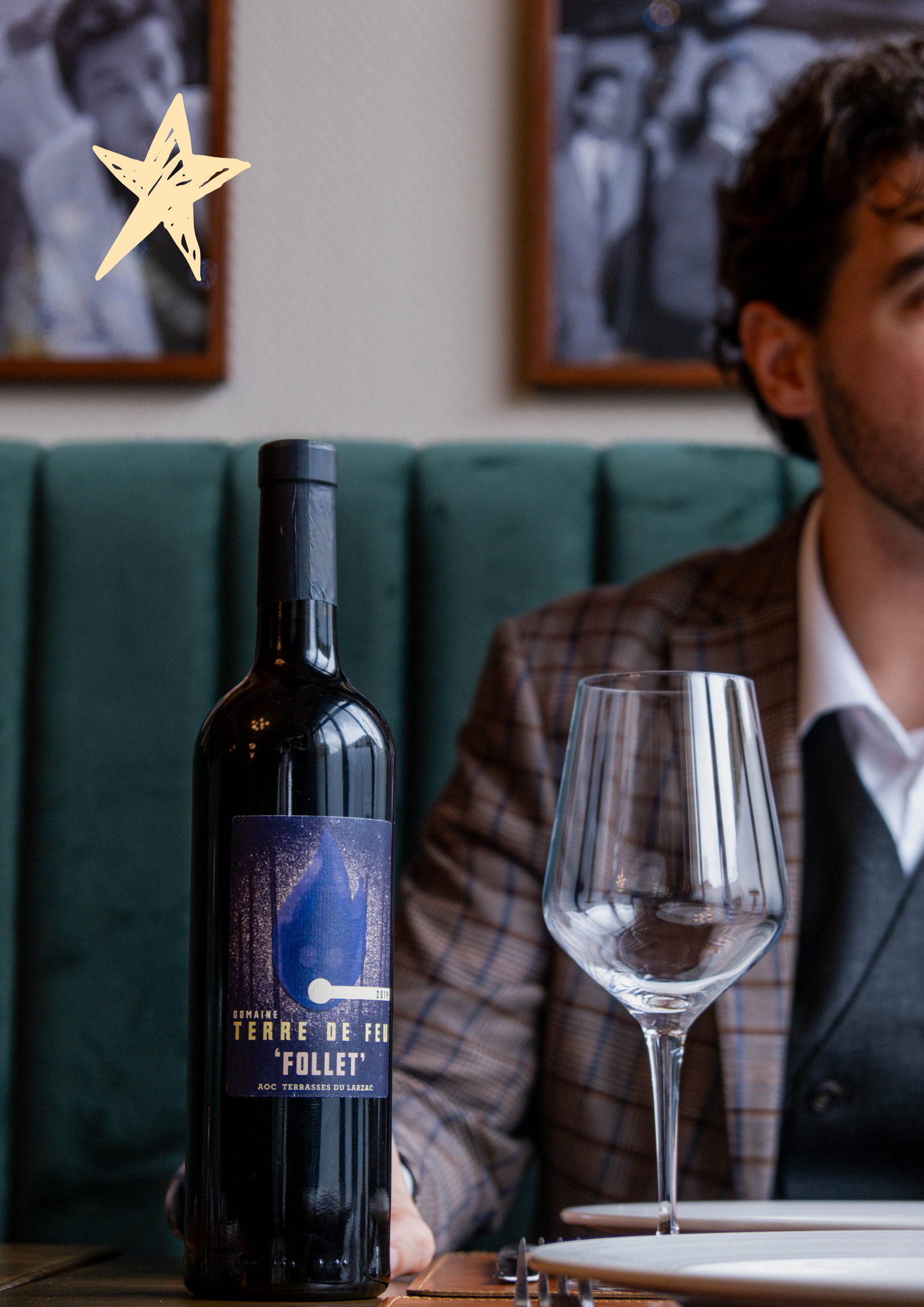 A bottle of red wine labeled "Terre de Feu 'Follet'" on a restaurant table with an empty wine glass beside it.  A wine taster is seen in the background. This is part of a Bonaventure Events experience.