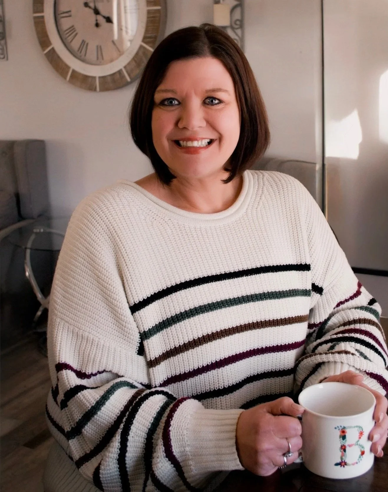 A woman with short brown hair smiling while holding a white mug with a floral letter 'B' on it, inside a kitchen with a wall clock behind her.