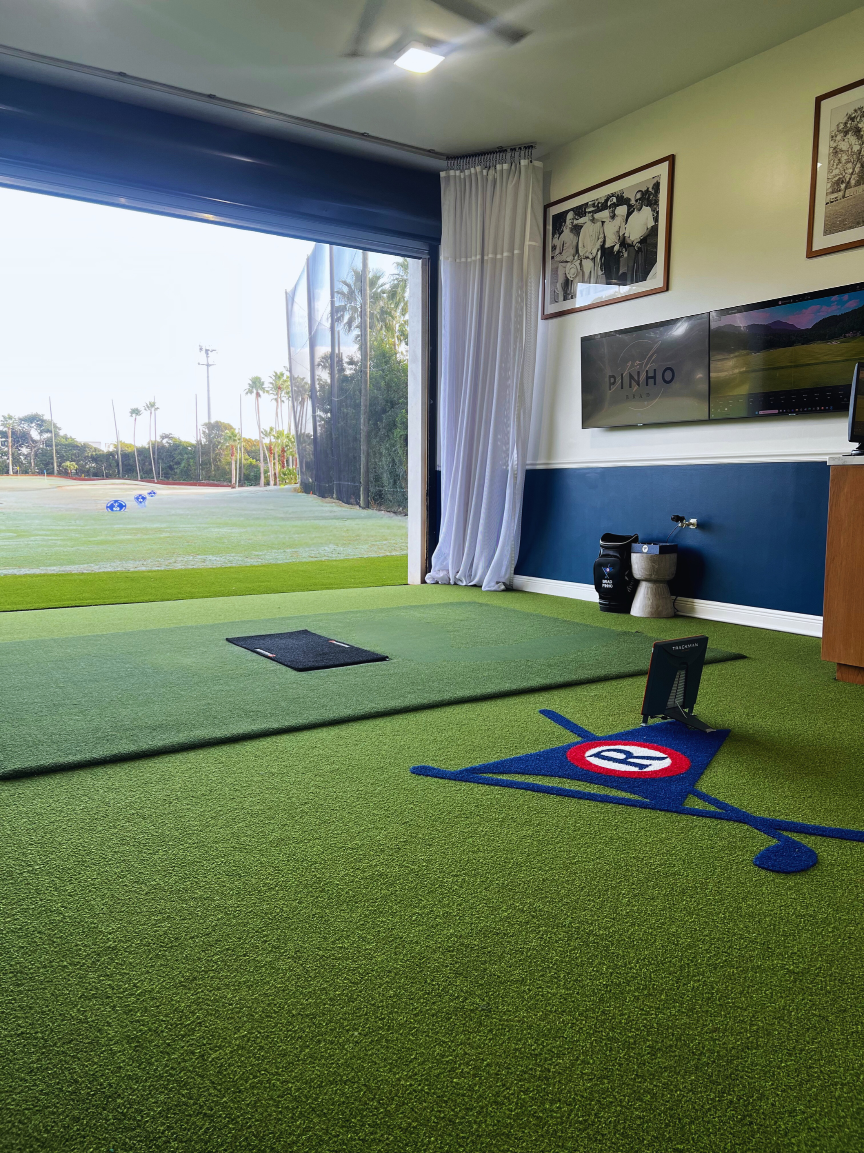 Indoor golf practice area with artificial turf, a mat, and a metal track training aid. Large window with white curtains showing outdoor scenery with palm trees and golf course. Wall decorated with framed black-and-white photos and a TV screen, with golf equipment and decor on the floor.