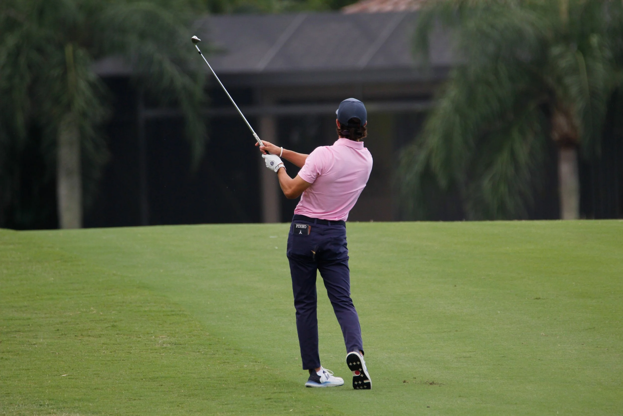 A golfer in a pink shirt, dark pants, and a cap taking a swing on a golf course.