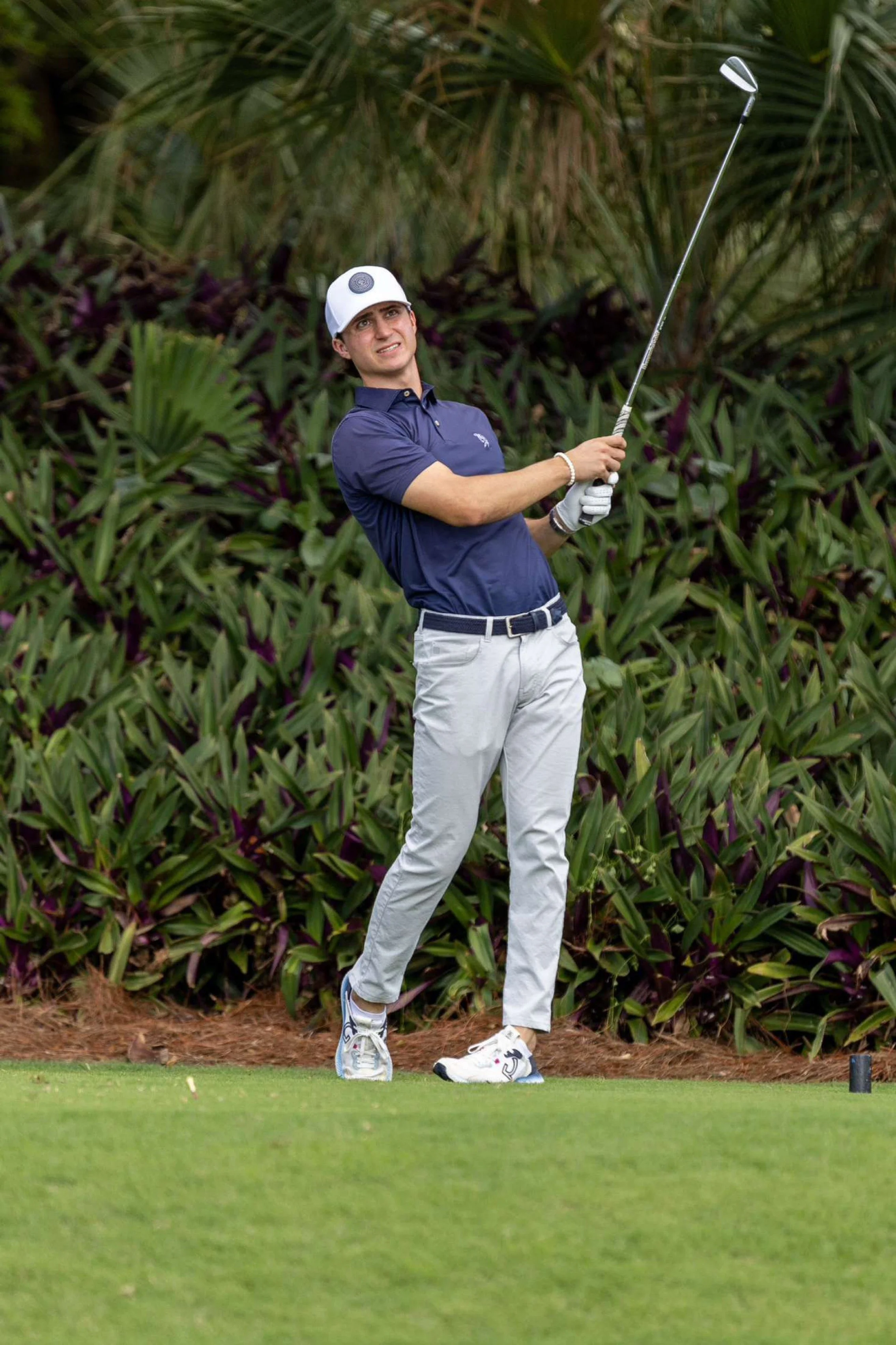 A young man playing golf on a course, holding a golf club after a swing, with trees and bushes in the background.