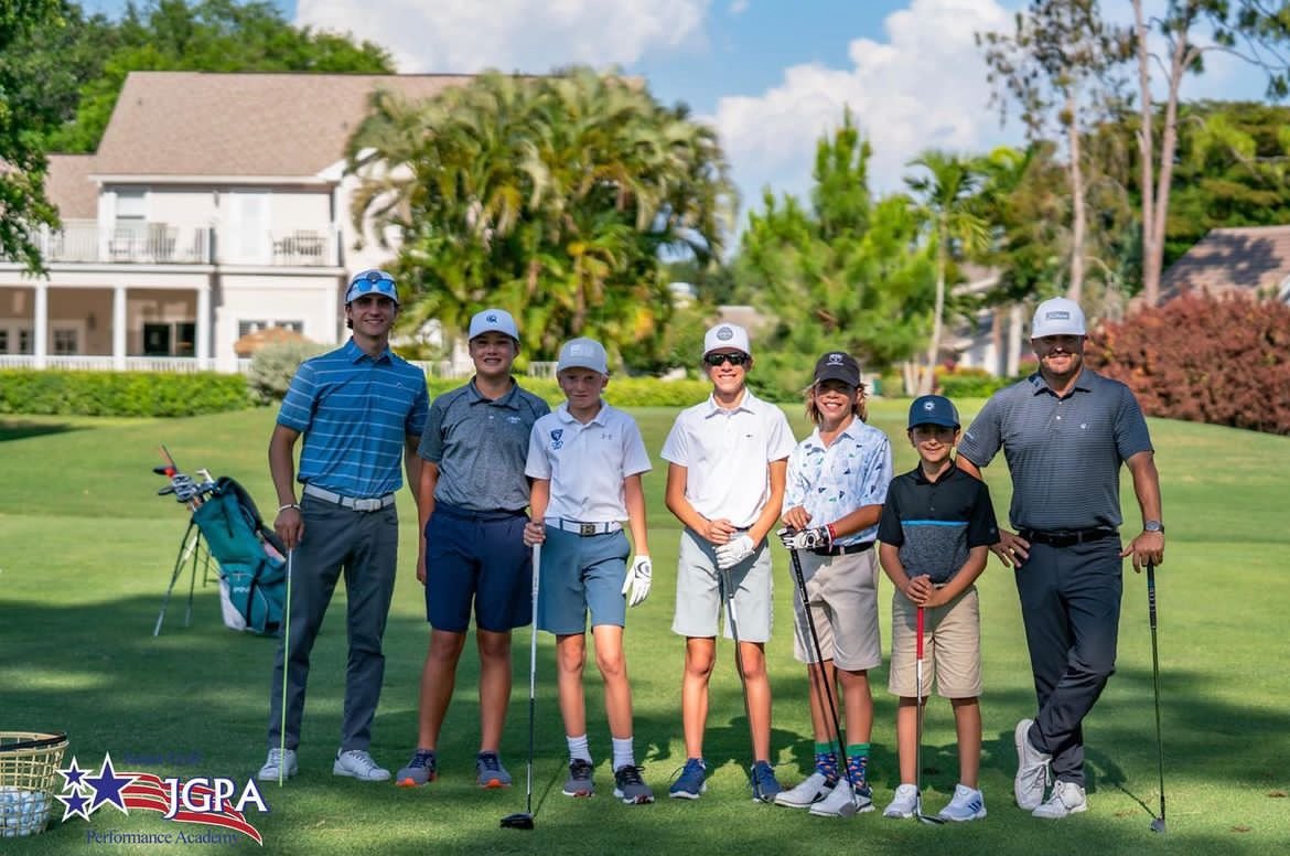 Group of eight people, including children and adults, standing on a golf course with golf clubs. They are dressed in golf attire, holding golf clubs, and smiling. There is a large house and trees in the background. The scene appears sunny and bright.