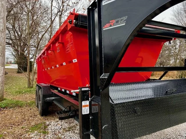 Red dump trailer parked on gravel with trees and a house in the background.