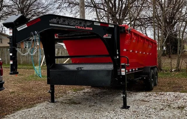 Red Horizon Trailmobile trailer with black hitch and support legs parked on gravel, trees and a wooden fence in the background.