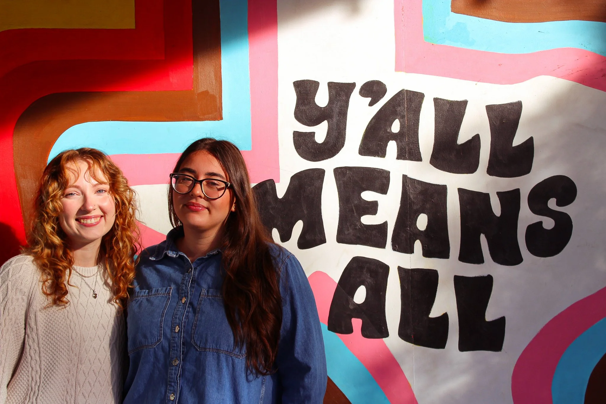 Two women standing in front of a colorful mural that says 'Y’all Means All' with vibrant abstract shapes.
