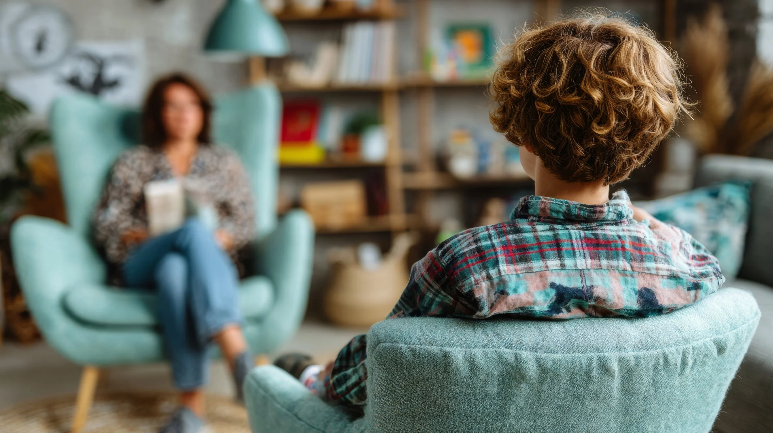 A child with curly hair sitting on a teal armchair, facing an adult woman with blurred features, sitting in a similar teal armchair in a cozy, decorated living room.