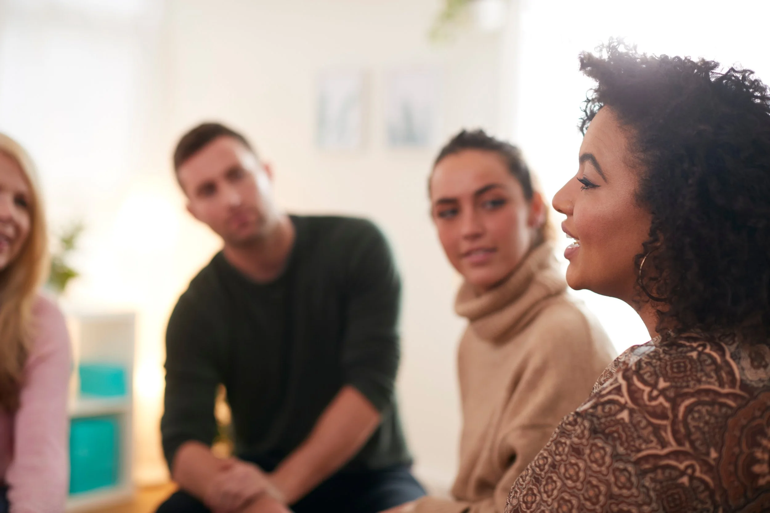 Group of four people sitting and talking indoors, with a woman in the foreground speaking and three others listening.