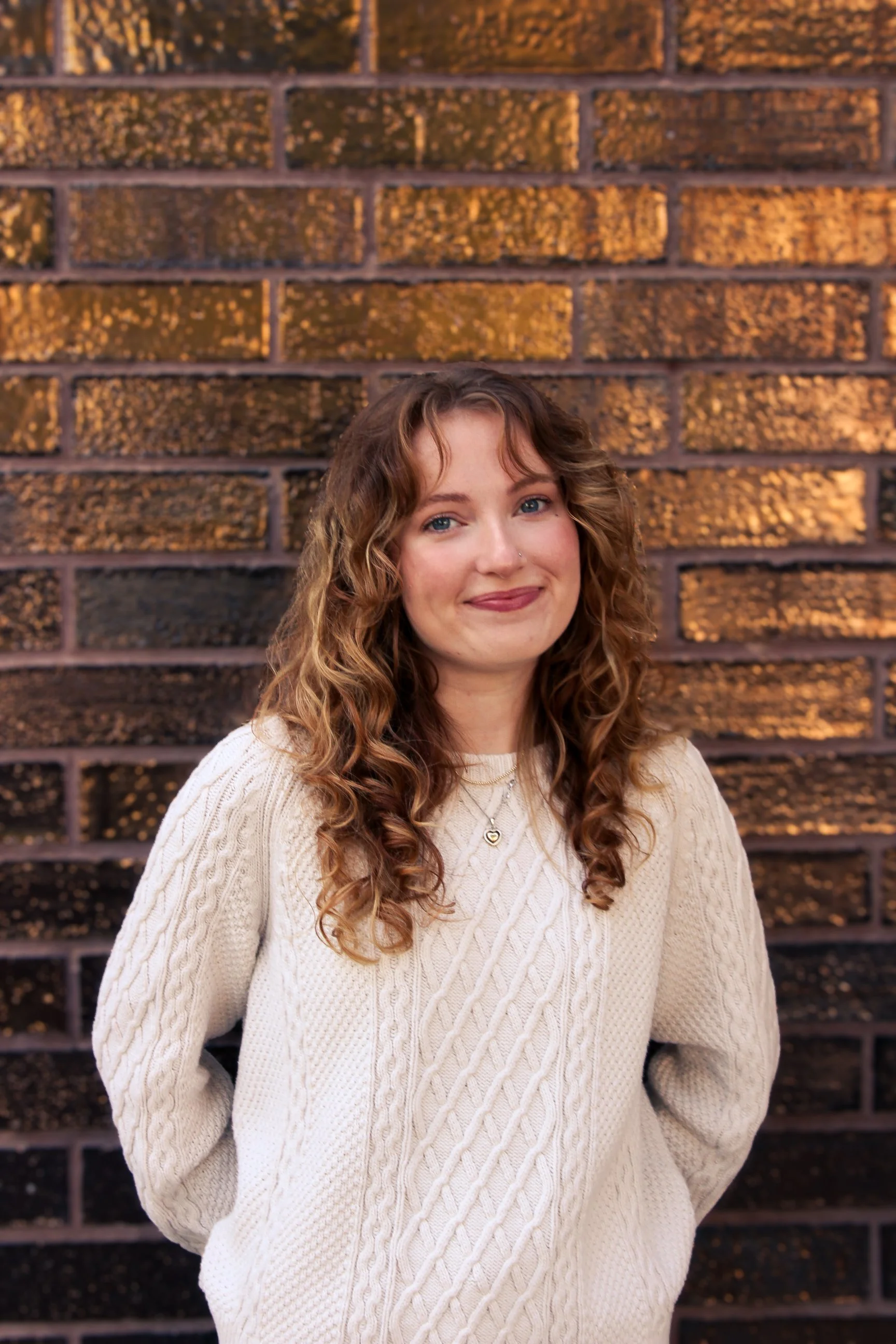 A young woman with long, curly brown hair, wearing a cream-colored cable-knit sweater and a gold necklace, standing confidently in front of a textured brick wall illuminated by warm lighting.