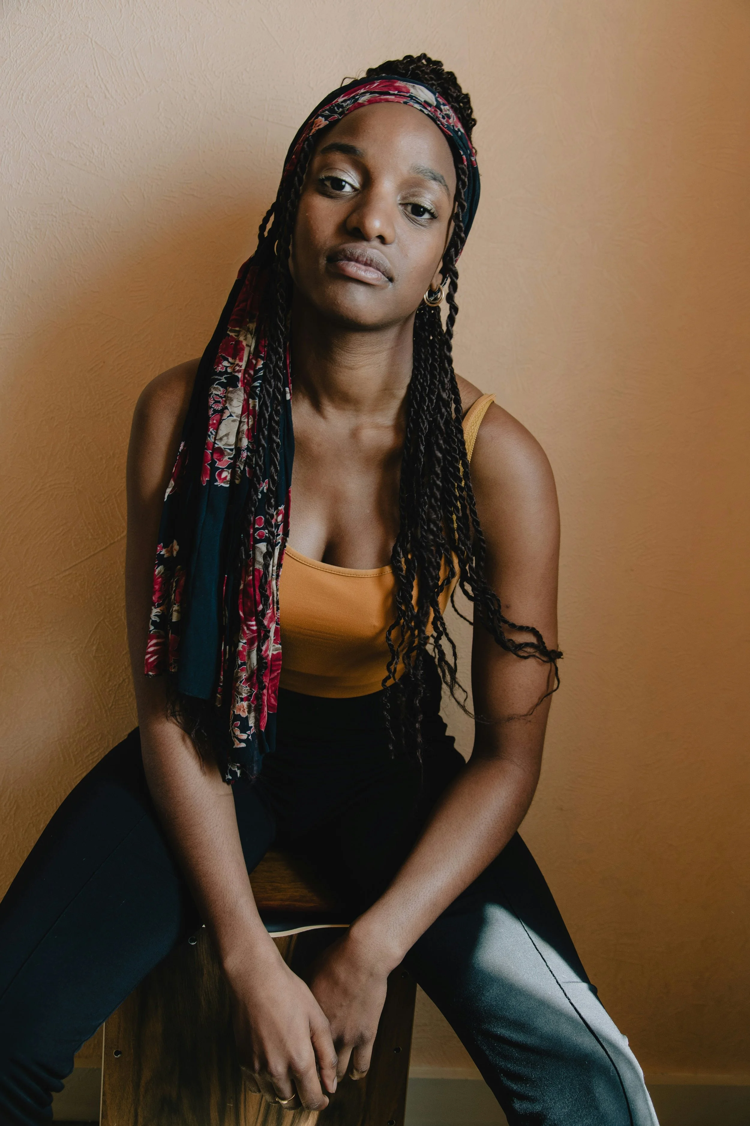 A young woman with dark skin and long braided hair sitting on a wooden stool against a beige wall. She wears a yellow tank top, dark pants with a light stripe, and a colorful scarf headband. Her expression is neutral.
