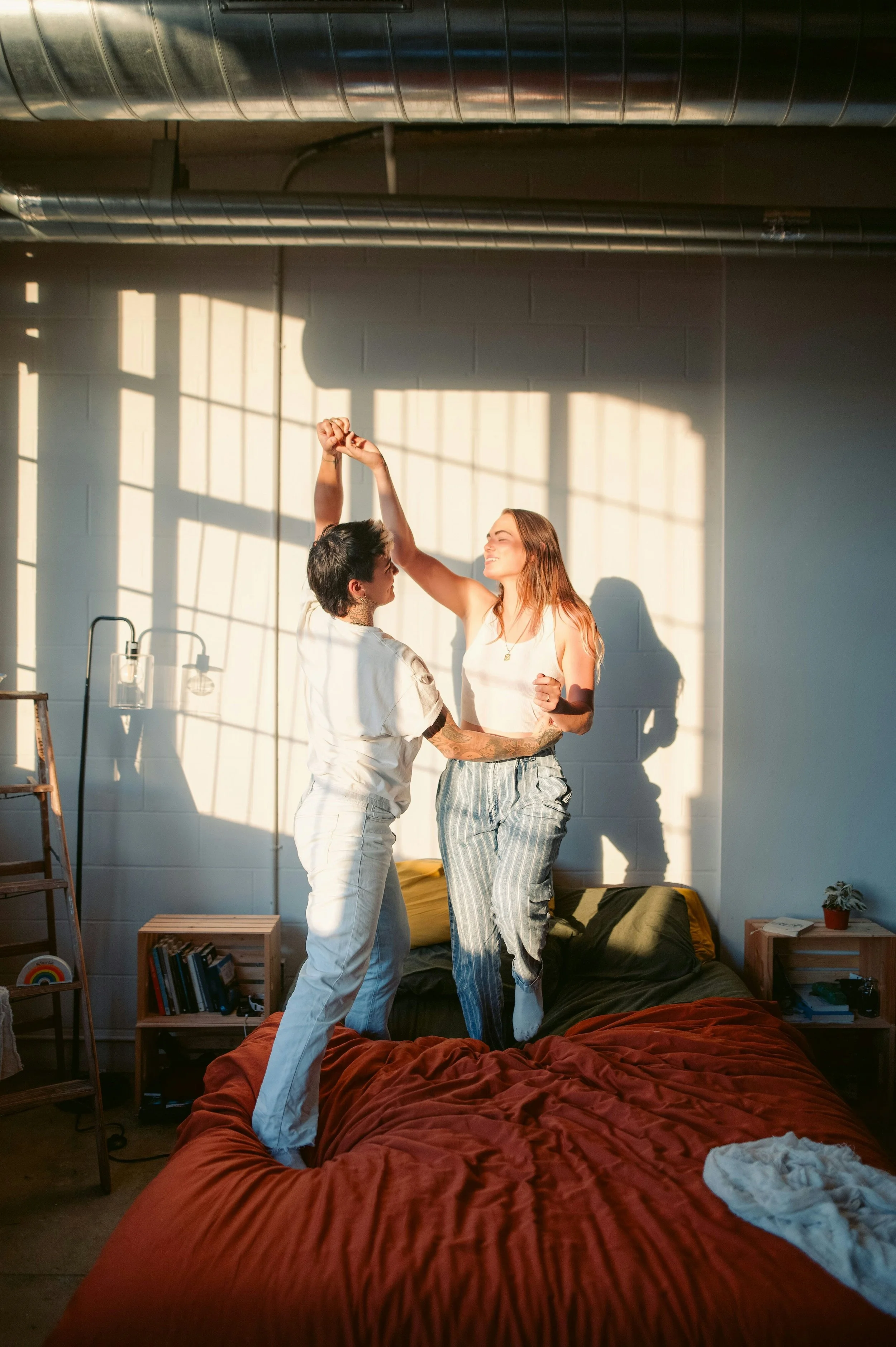 Two women dancing on a bed in a sunlit bedroom, with one woman lifting her arm and the other smiling.