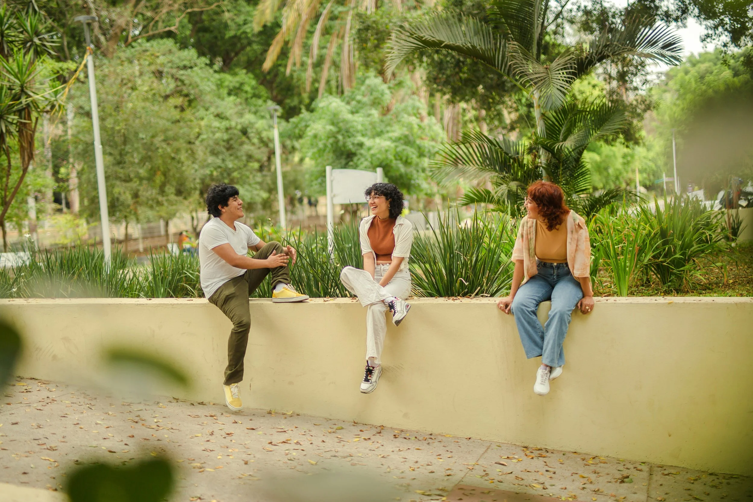 Three friends sitting on a low wall in a lush green park, smiling and engaging in conversation.