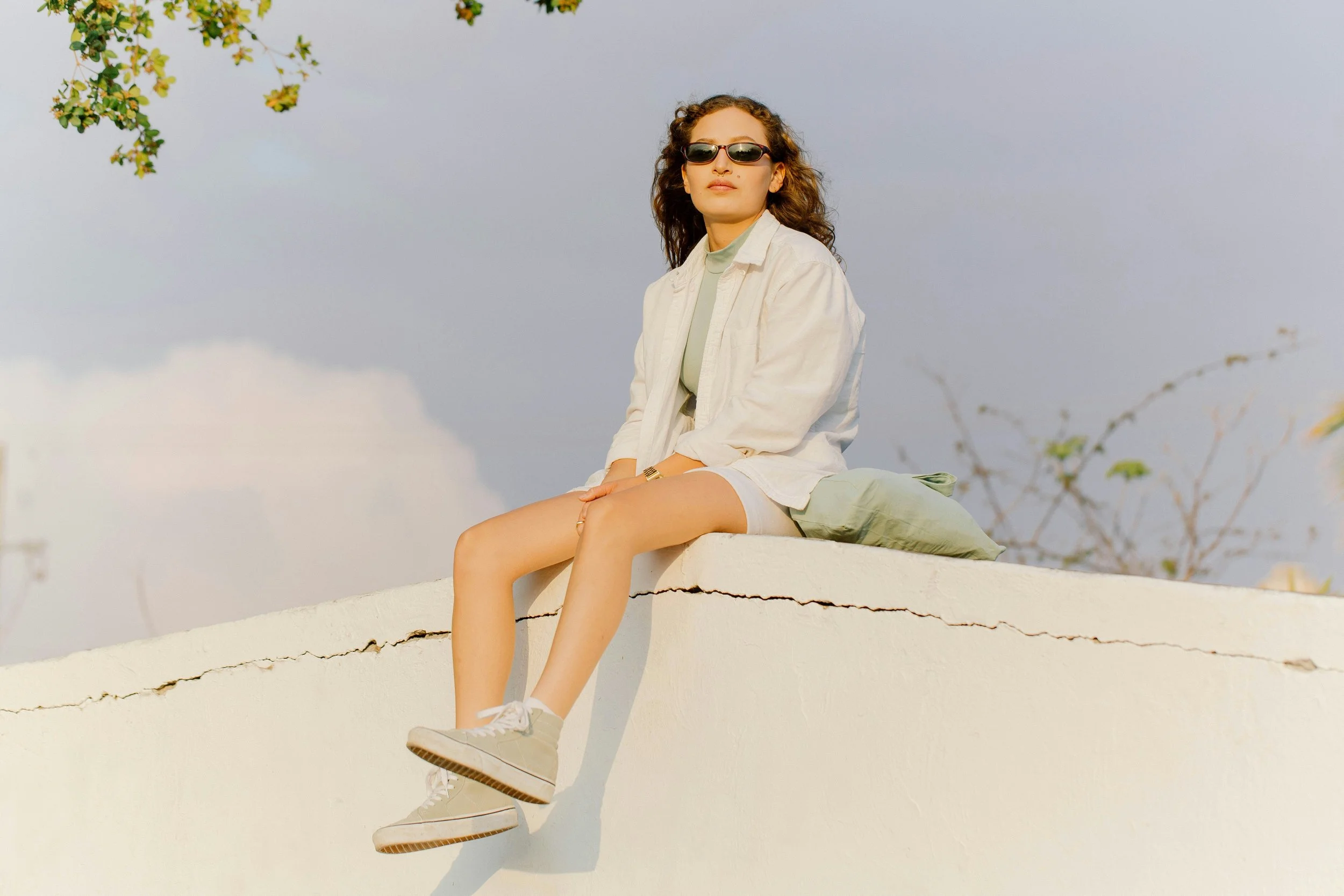 A young woman with curly hair and sunglasses sitting on a white wall outdoors, wearing a light-colored jacket, shorts, sneakers, and a watch, with a sky and some trees in the background.