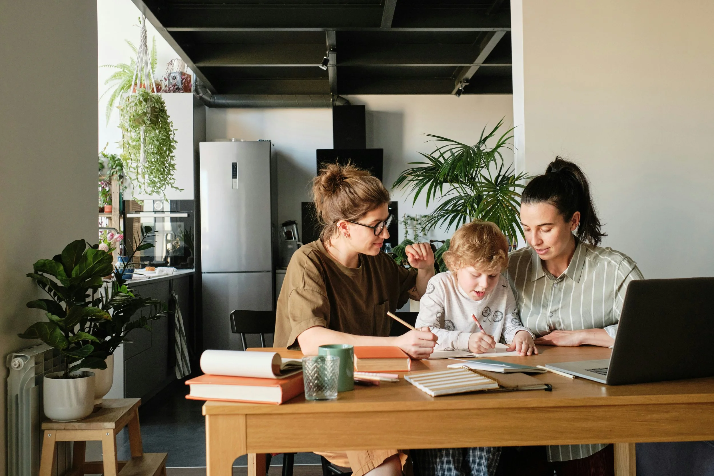 Three people, two women and a young boy, sitting at a wooden table working together with notebooks, pencils, and a laptop in a cozy, plant-filled kitchen or dining area.