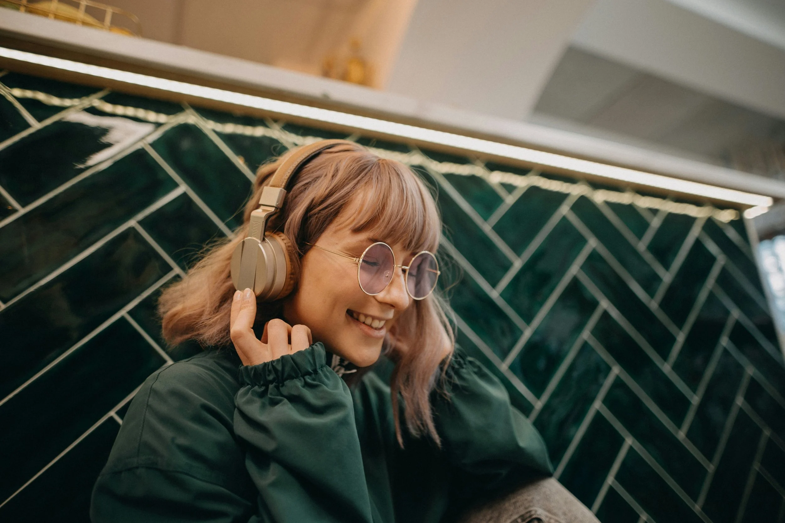 A young woman with blonde hair, wearing round glasses and beige headphones, smiling and leaning against a green tiled wall.