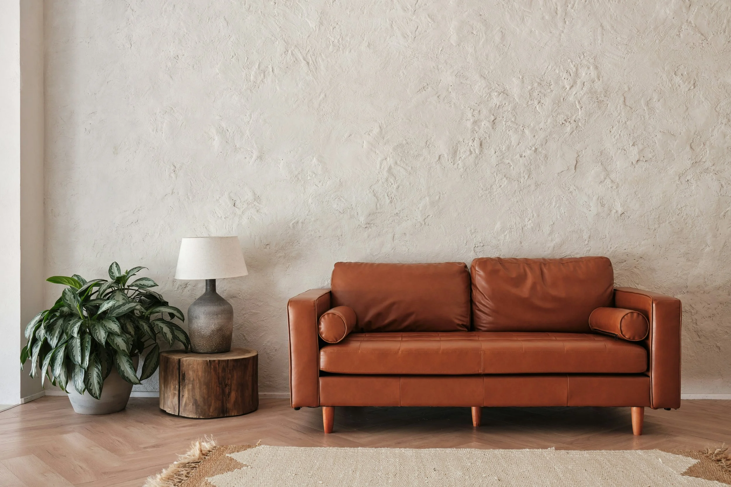 Living room with a brown leather sofa, a wooden side table, a table lamp, and a potted plant against a textured cream wall.