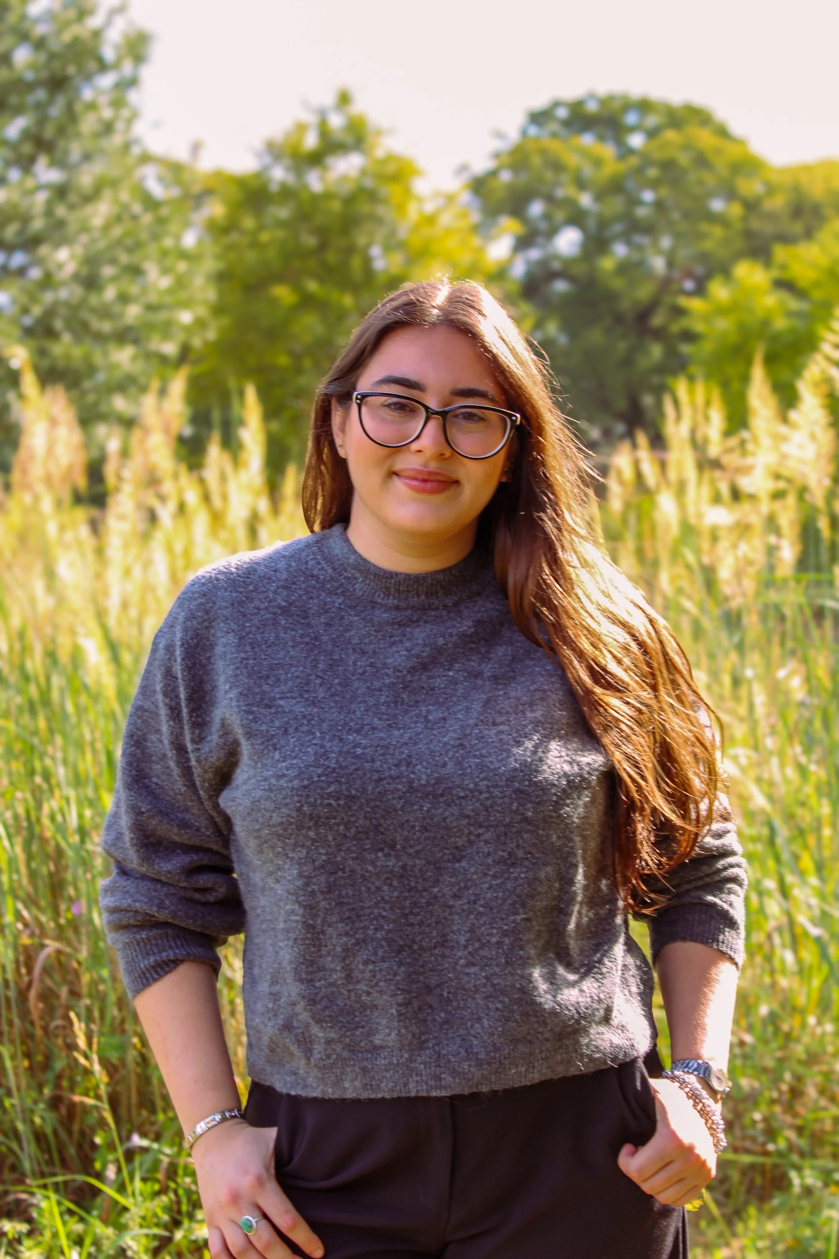 Young woman with glasses and long brown hair standing outdoors in a field of tall grass and trees, smiling at the camera on a sunny day.