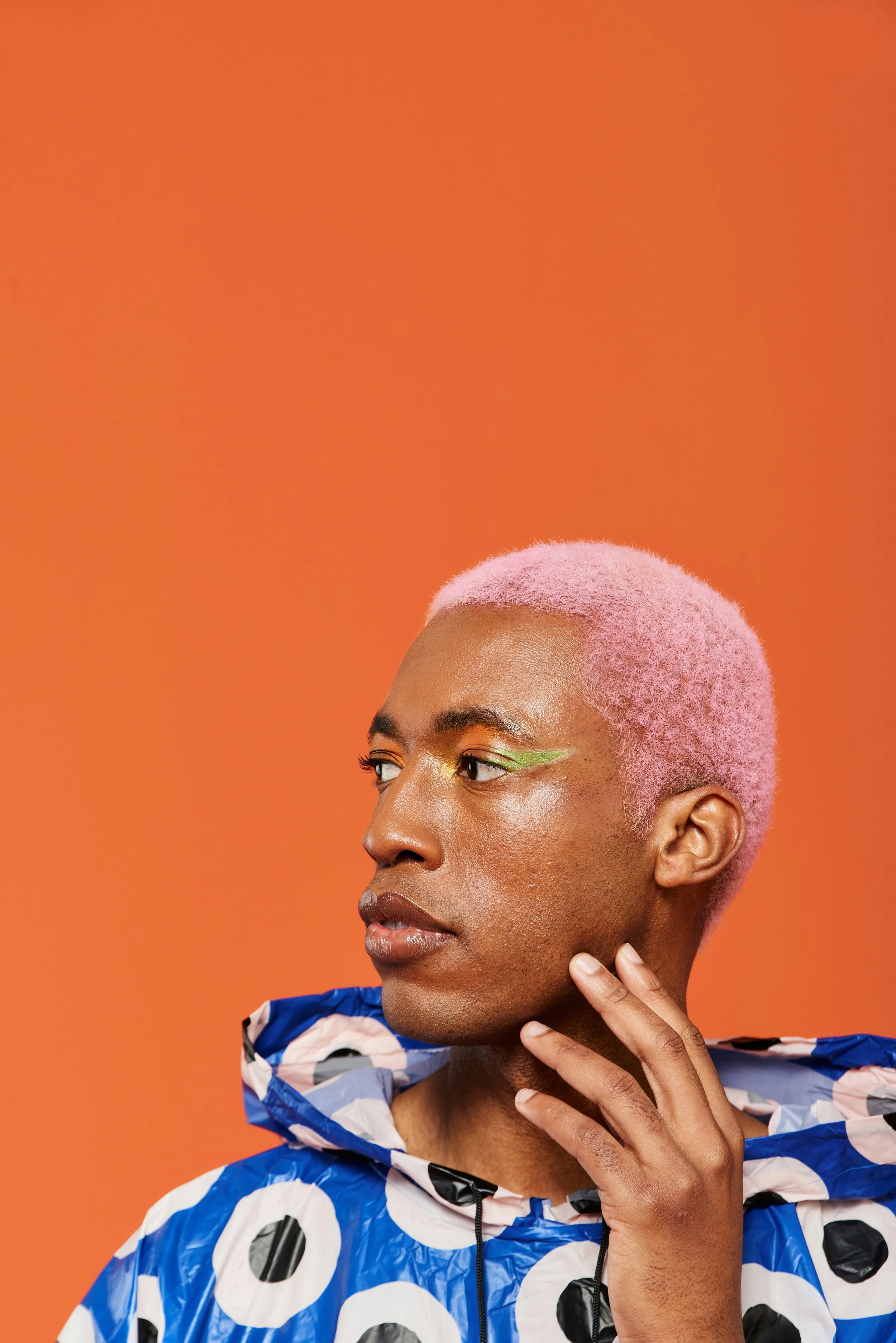 Portrait of a person with pink hair, artistic green eye makeup, wearing a blue and white patterned jacket, against an orange background.