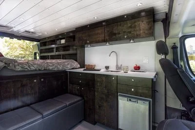 Interior of a converted camper van with a kitchen area featuring dark wood cabinets, a small sink, and a compact fridge, with a bed at the back and seating area in the front.