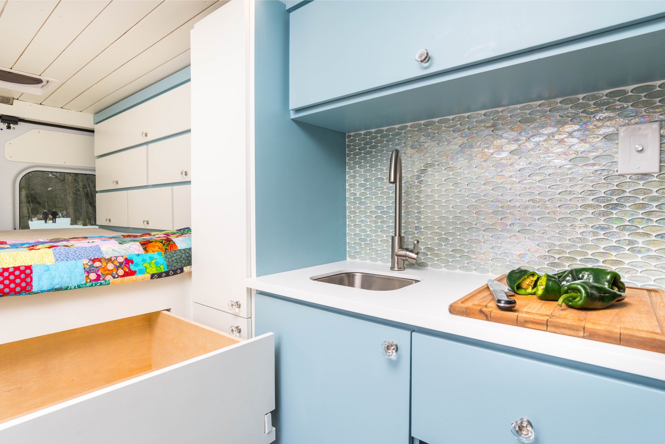Interior of a tiny home with a small kitchen featuring blue cabinets, a white countertop, a tile backsplash, and a wooden cutting board with green bell peppers. In the background, a bed with a colorful patchwork quilt is visible.