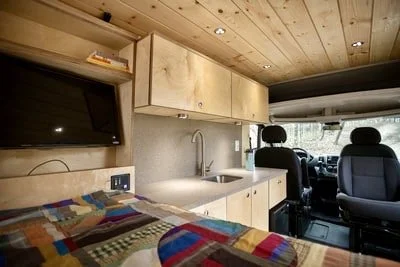 Small kitchen area with light wooden cabinets, a sink, a faucet, a countertop, a flat-screen TV on the wall, and two front seats in a vehicle