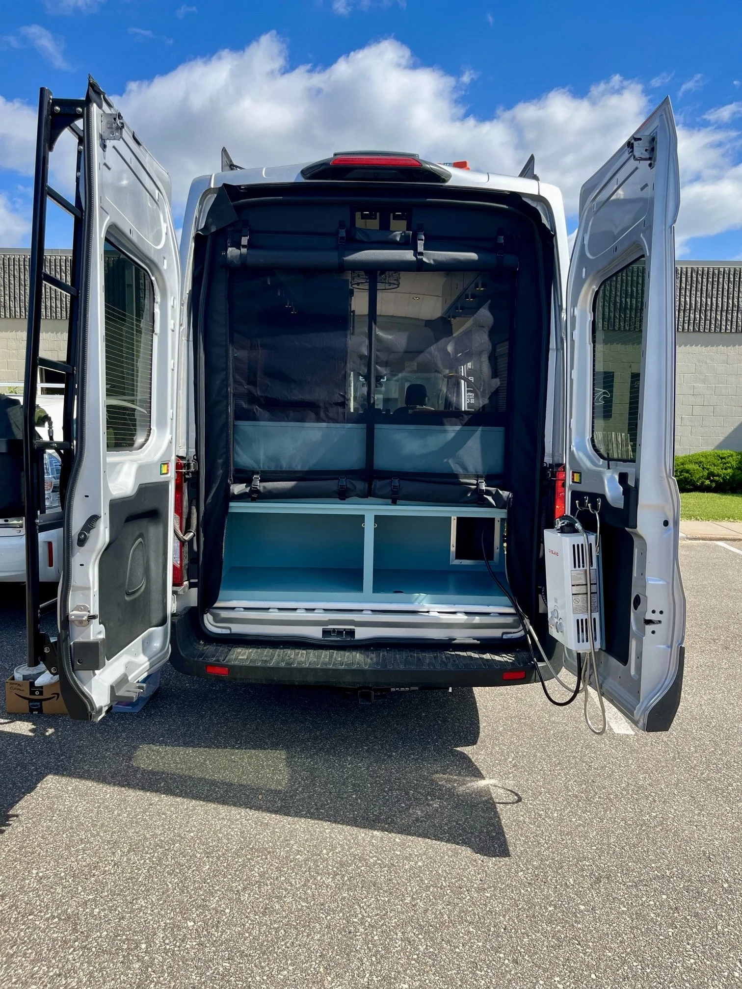 Inside the back of a white van, showing a black screened pet barrier, some blue storage compartments, and electrical equipment, with the van open on a parking lot under a partly cloudy sky.