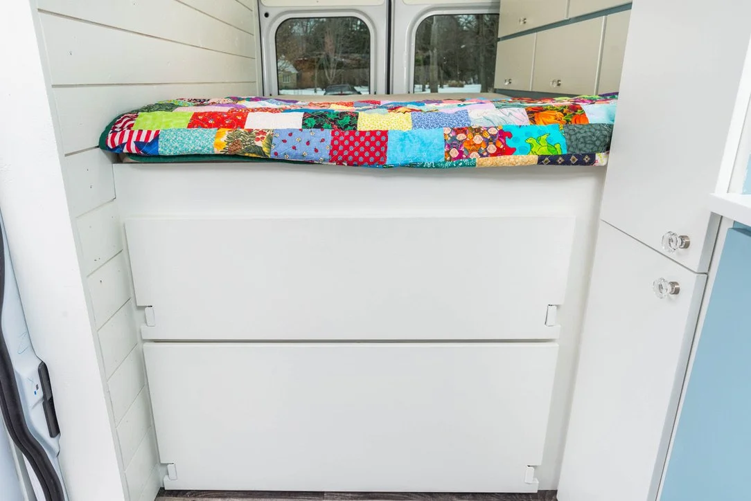 A hidden bed in a camper van with a colorful patchwork quilt on top, situated above storage drawers.