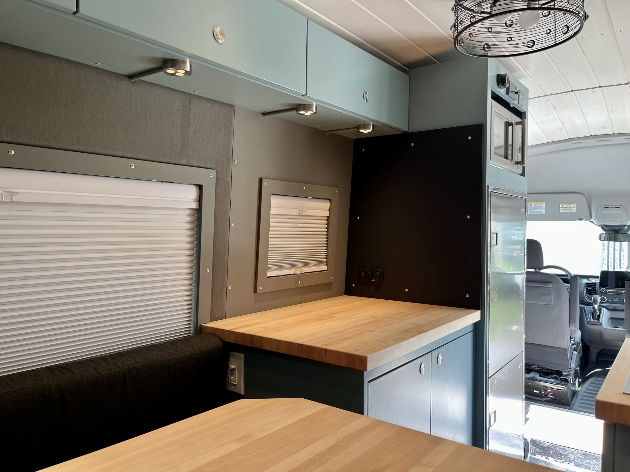 Interior of a camper van showing a wooden countertop, small window with a roller shade, gray and teal cabinetry, black wall panel with switches, refrigerator, and driver’s seat at the front.