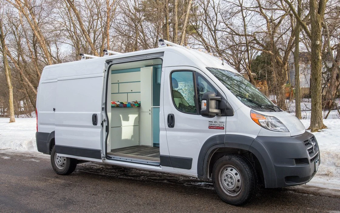 White cargo van with side door open, showing shelves inside, parked on snowy roadside with leafless trees in background.