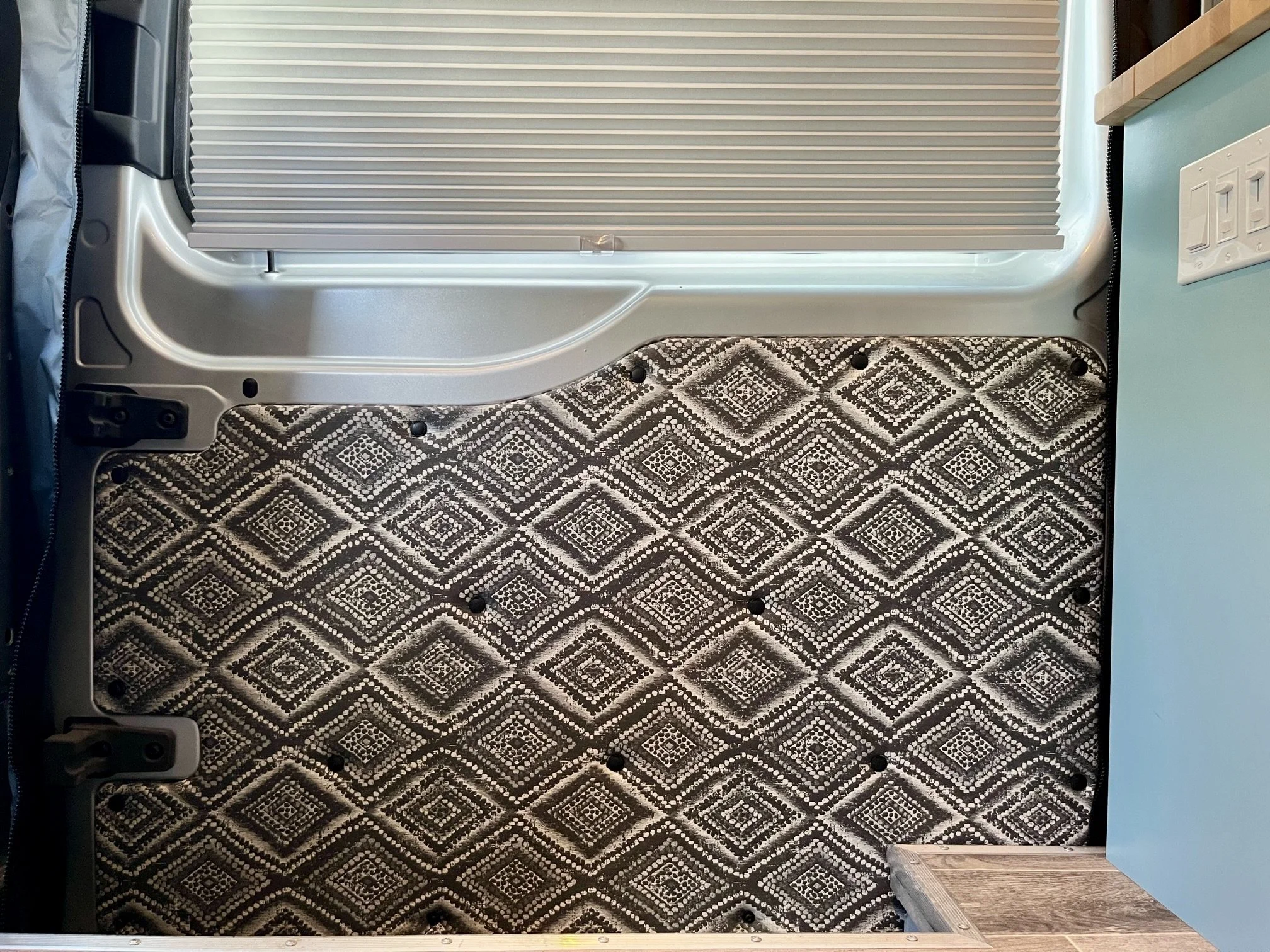 Interior of a van with a patterned black, white, and grey insulation panel on the wall, a light blue wall, an electrical outlet, and a small wooden step at the bottom right.