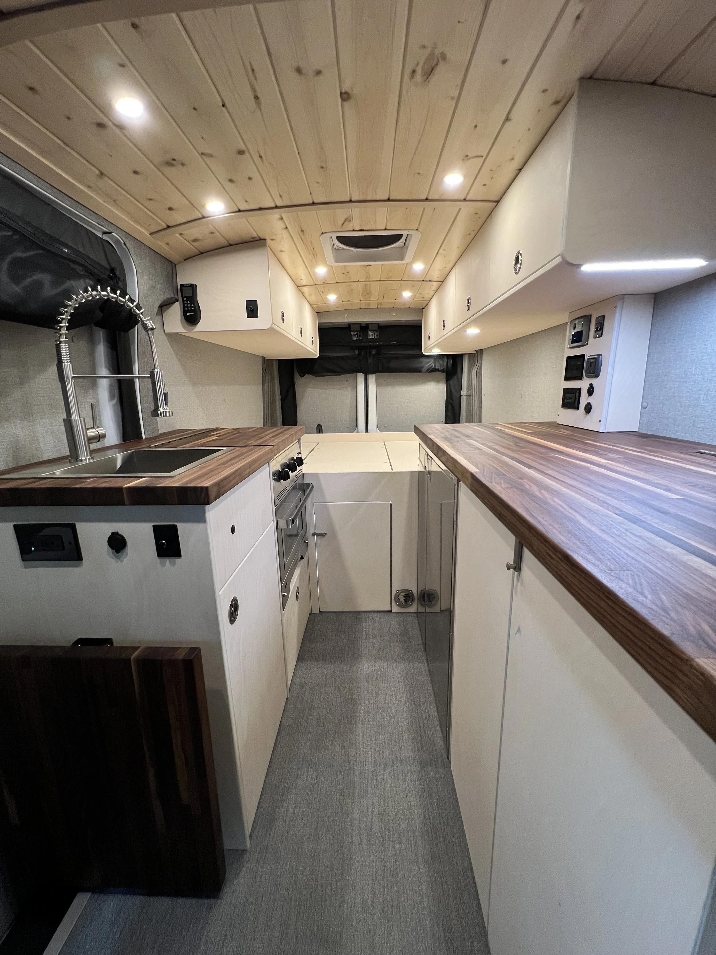 Interior of a compact camper van kitchen with a wooden ceiling, white cabinets, a small sink with a flexible faucet, a stove, and a large wooden countertop on the right side.