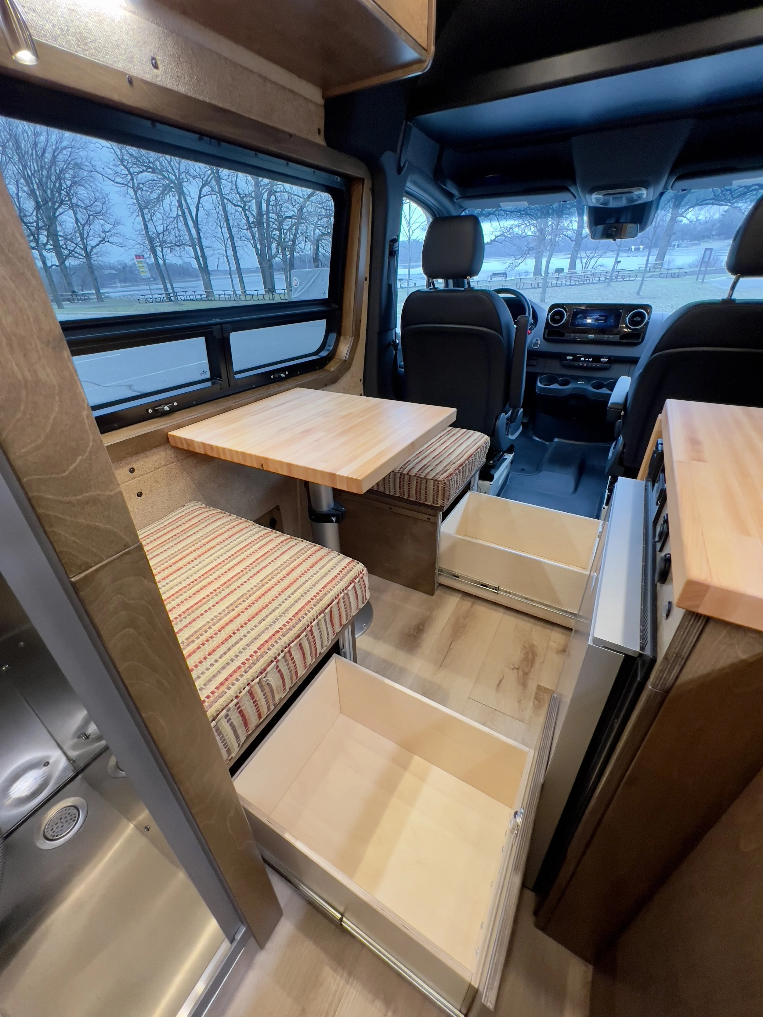 Interior of a camper van with wooden cabinetry, striped cushions, a small table, and open storage drawers.