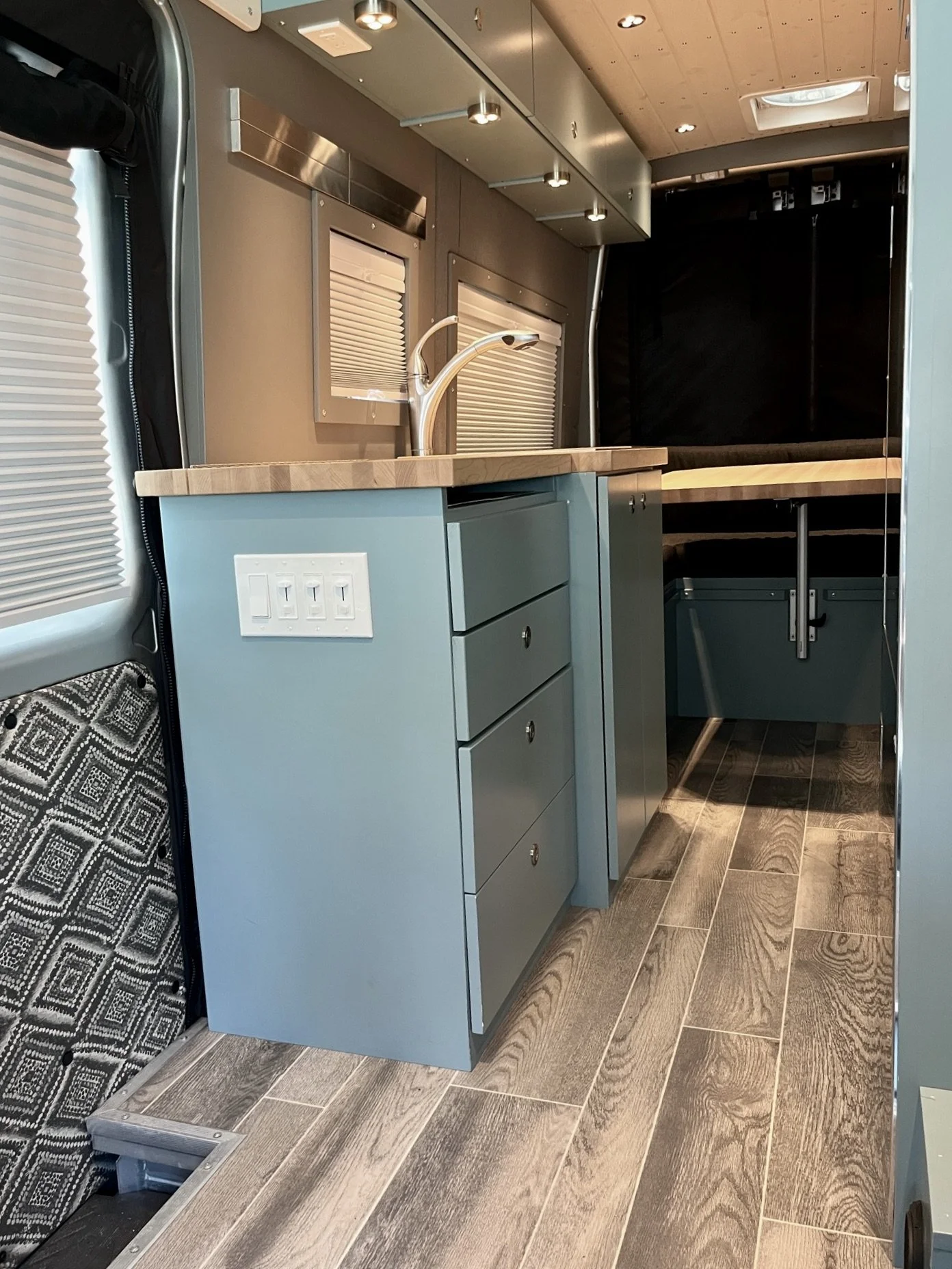 Interior of a compact, modern kitchen area inside a vehicle, featuring a blue cabinet with three drawers, a wooden countertop with a sink, a silver faucet, and overhead lighting. There are small windows with blinds and wood paneling on the ceiling.