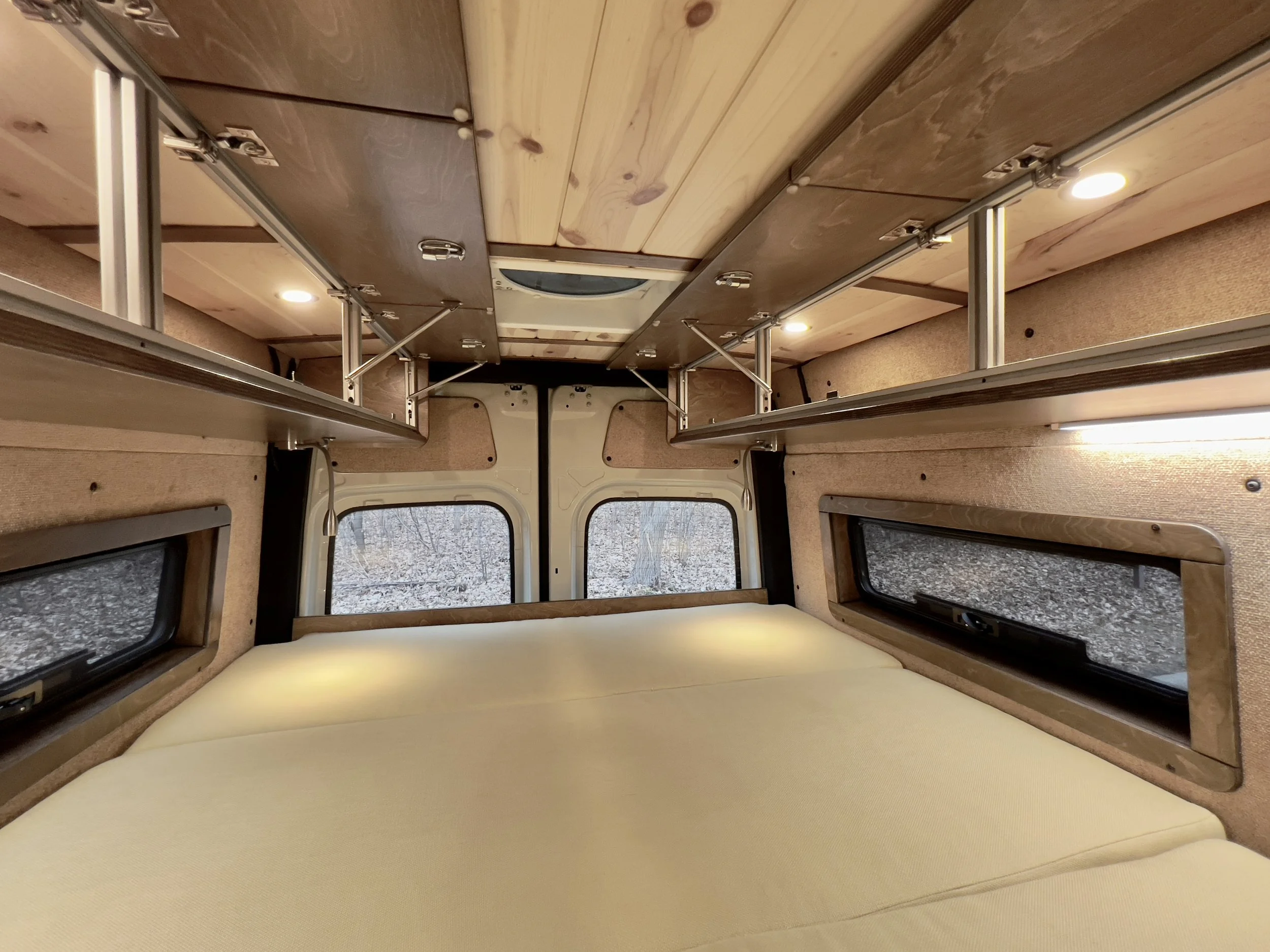 Interior of a camper van with a memory foam bed, small windows, and overhead storage shelves.