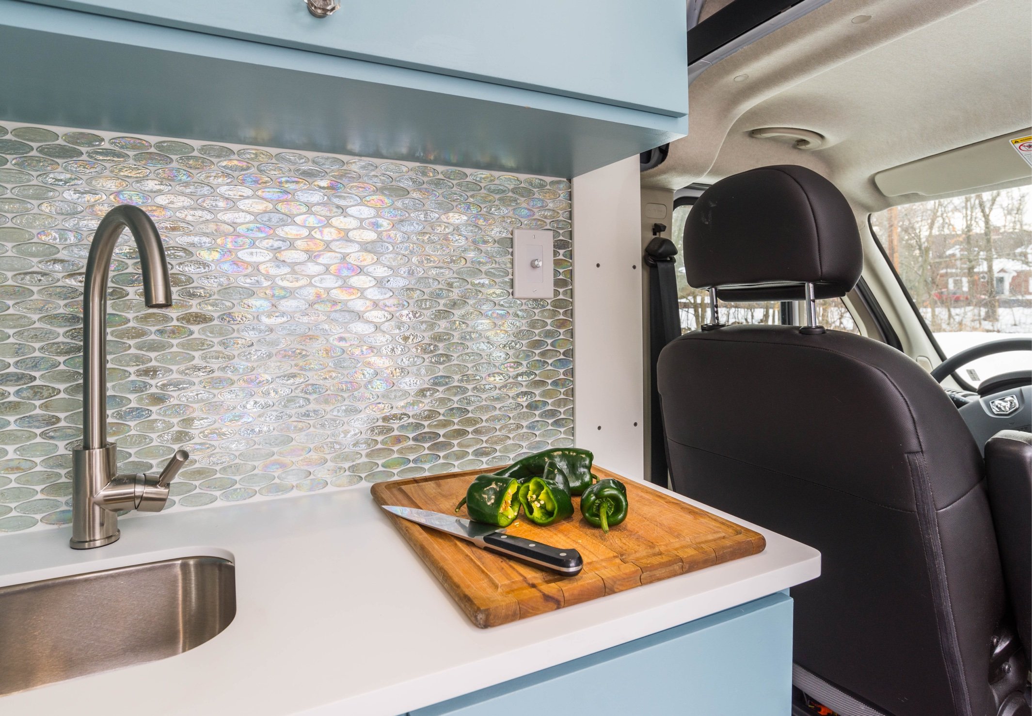 Inside a camper van kitchen, a cutting board with green bell peppers and a knife on it. The kitchen has a mosaic tile backsplash, a stainless steel sink with a modern faucet, and light blue cabinetry. The view shows the front seats and windshield of the van, with a snowy landscape outside.