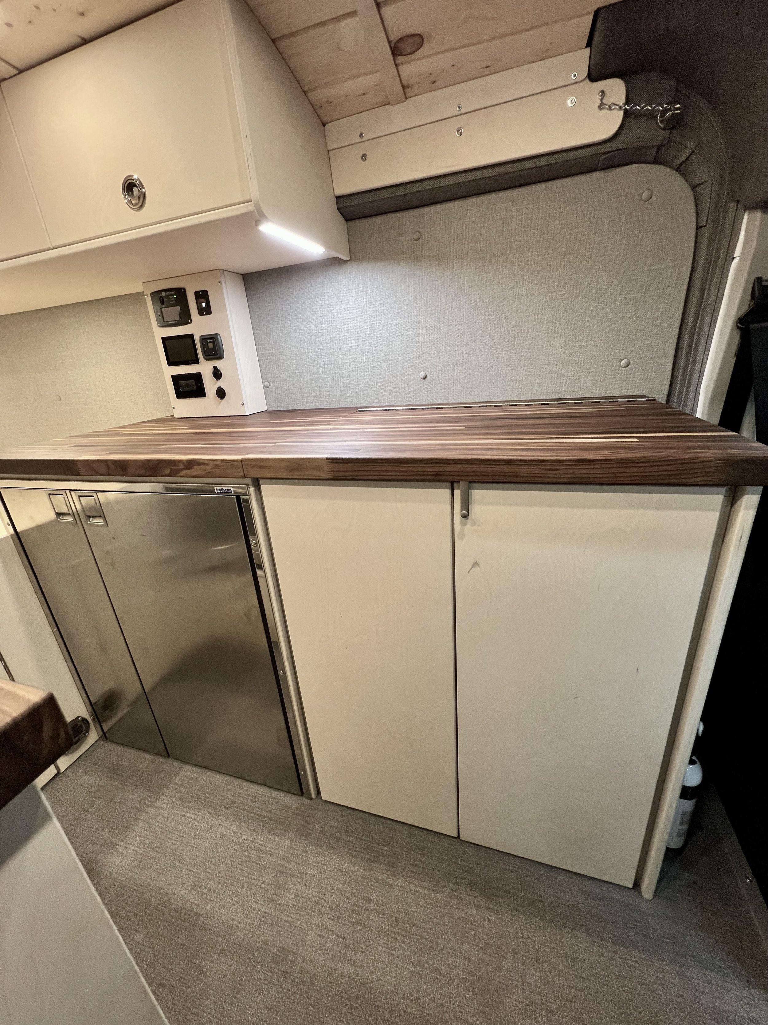 Part of a compact kitchen with beige cabinets, a wooden countertop, and a small stainless steel refrigerator, set inside a camper or tiny house.