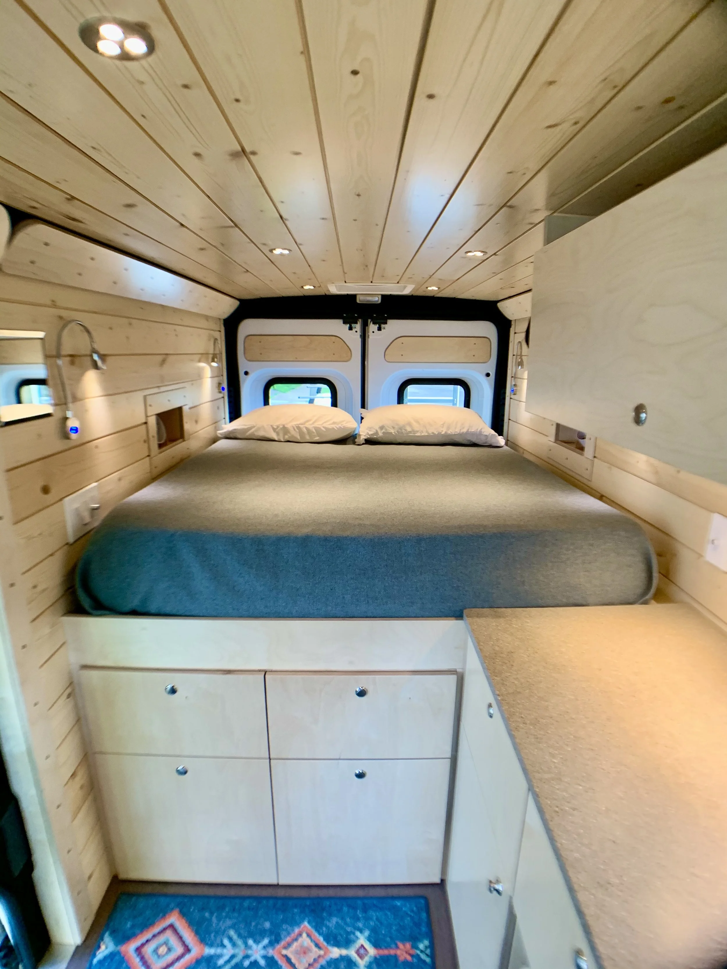 Interior of a small camper van with wooden paneling, a bed with gray bedding and pillows, small windows at the head of the bed, and built-in cabinets and drawers in light wood.