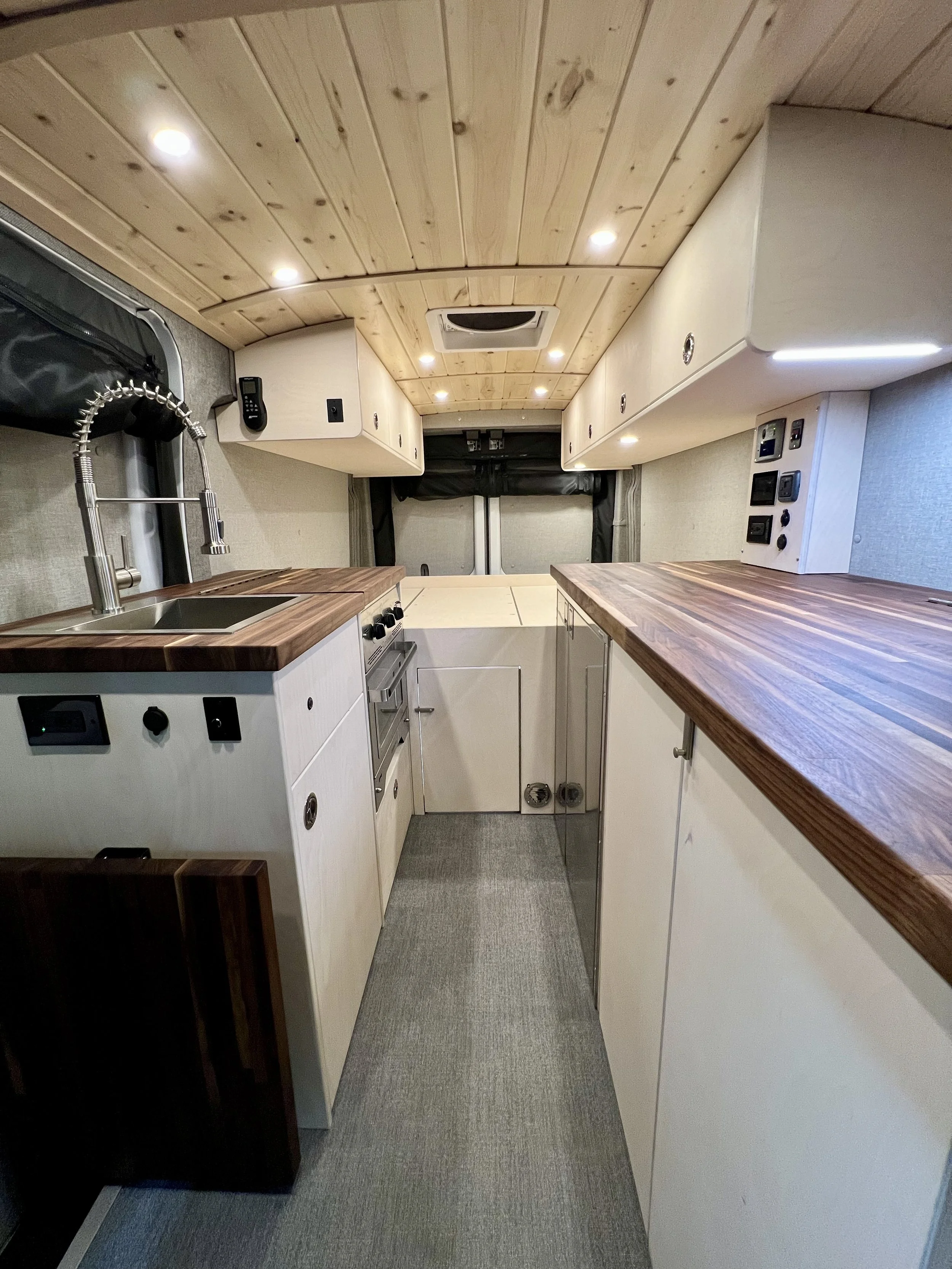 Interior of a compact camper van kitchen with wooden ceiling, white cabinetry, and wooden countertops. Includes a small sink with a high-arc faucet, a stove with oven, and various electrical outlets and switches.