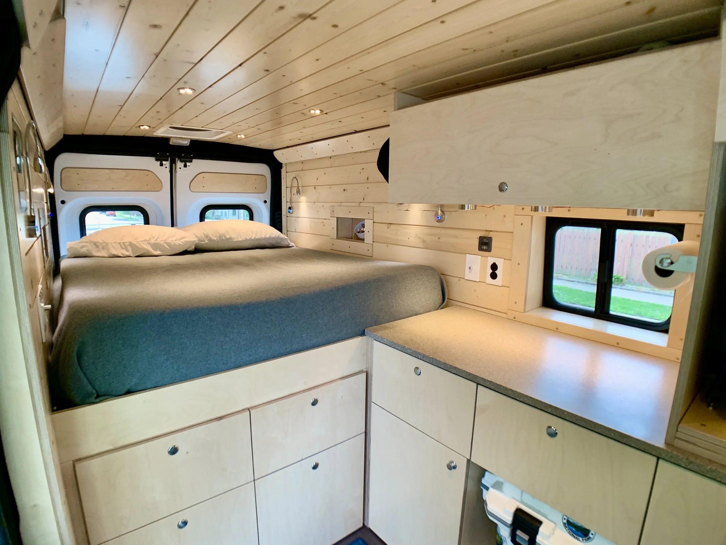 Interior of a small camper van with a bed, wooden cabinets, a window, and modern lighting.