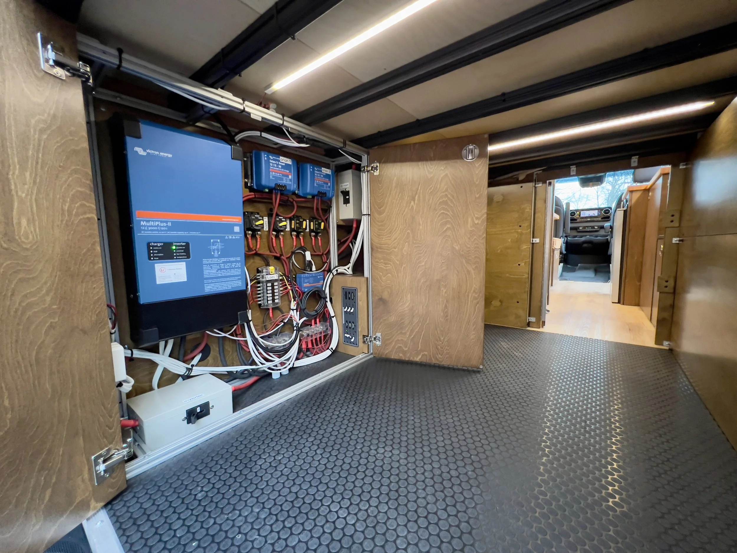 Electrical control system inside a wooden cabinet in a recreational vehicle, with wires, circuit breakers, and a large blue inverter or charger unit. The cabinet is open, revealing the equipment, and the RV's driver cabin is visible in the background.