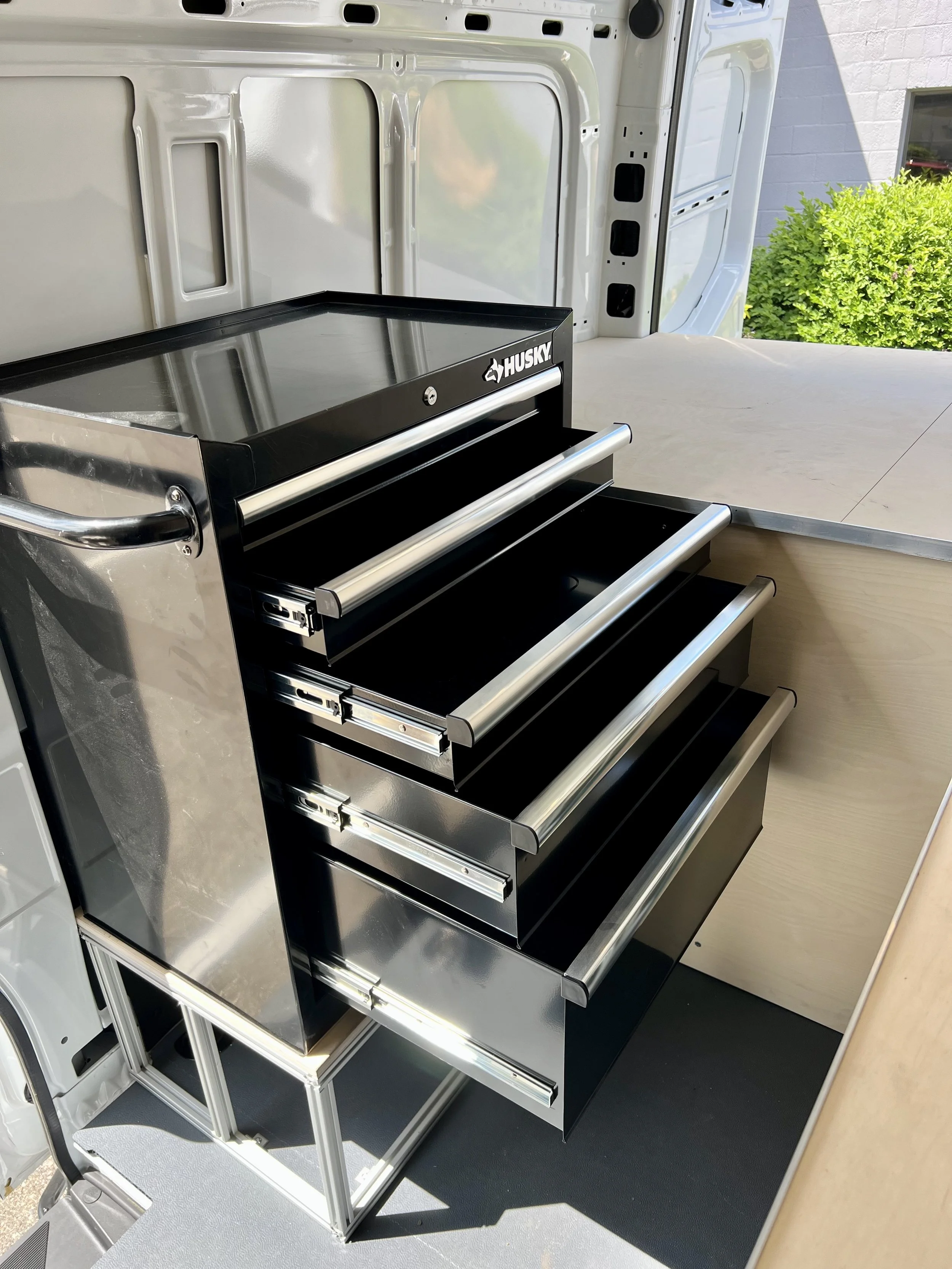 A black Husky tool chest with four drawers, situated inside a van with open back doors, next to a wooden work surface, outdoors with green bushes in the background.