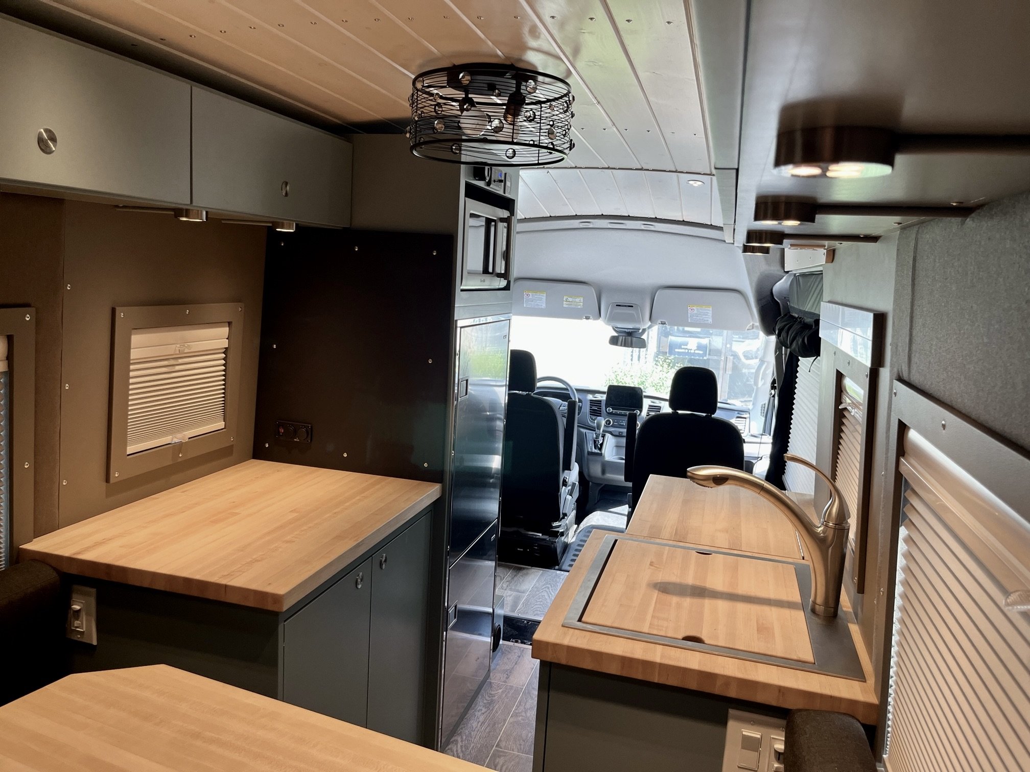 Interior of a camper van with a wooden countertop, kitchen sink, cabinets, and a view of the driver's cab. The space is compact and well-organized with natural light coming in from the front.