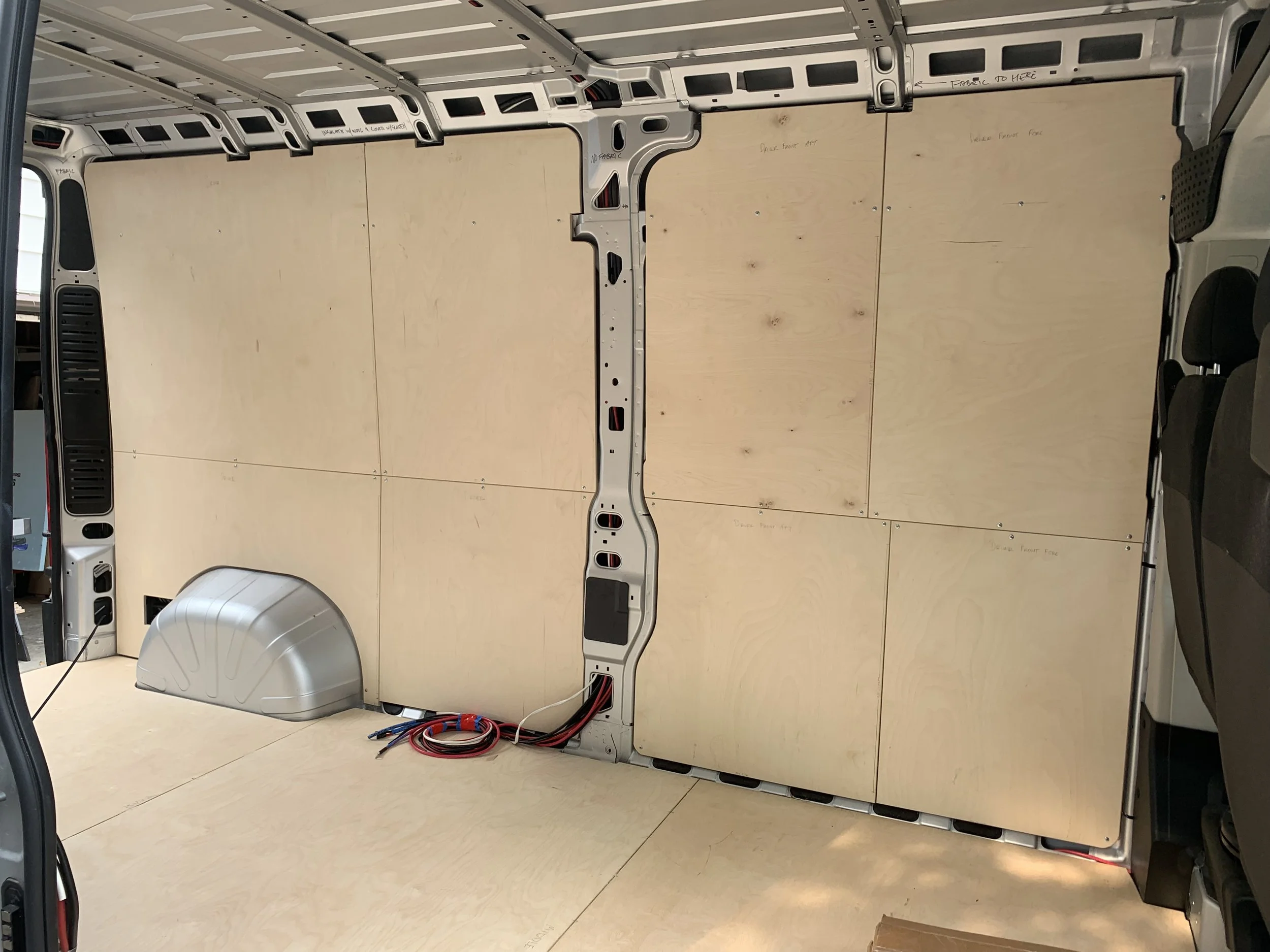 Interior of a vehicle with plywood panels attached to the walls and floor, exposed metal framework, wiring on the floor, and part of a silver metal component near the wheel well.