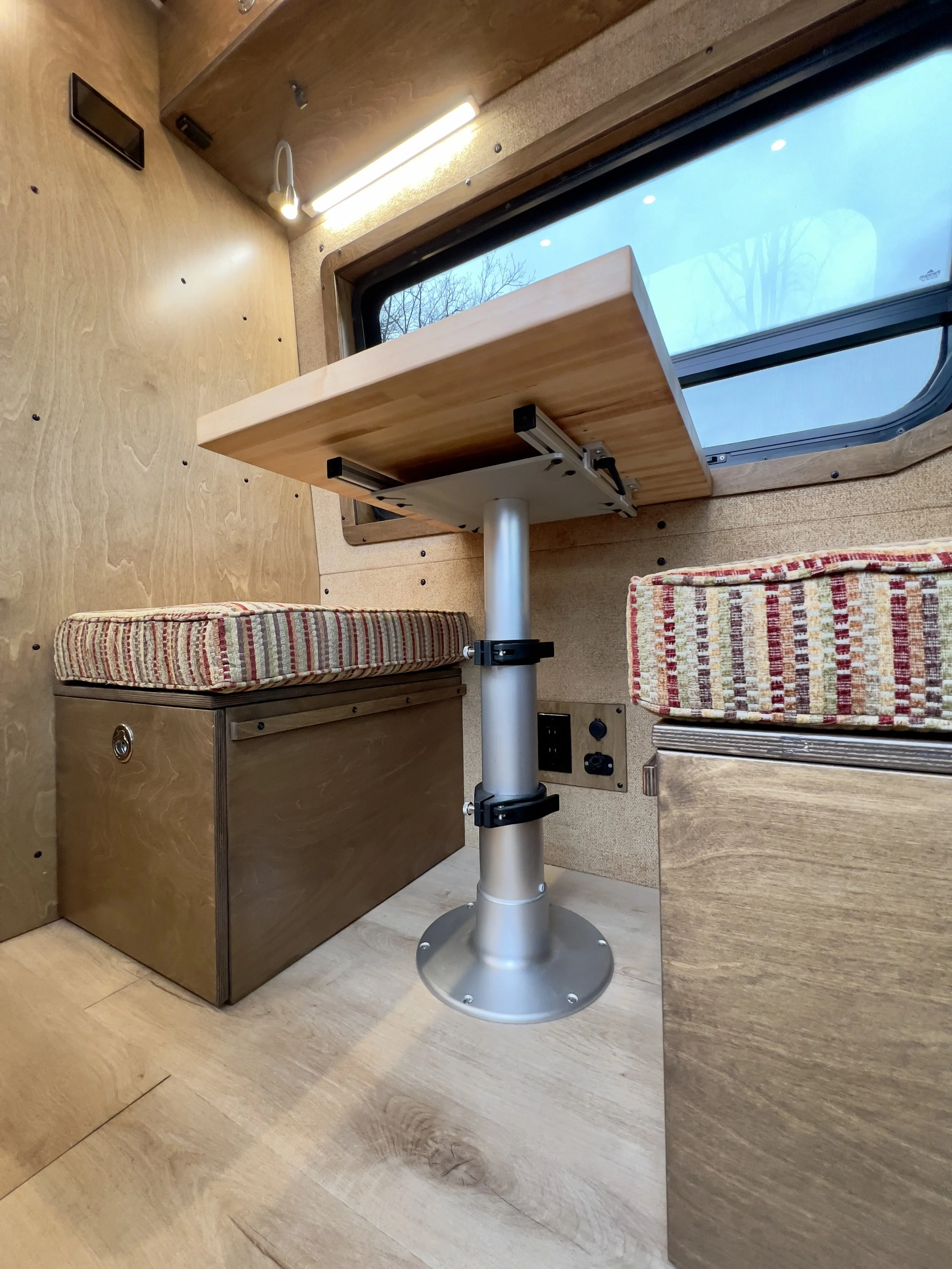 Interior of a compact living space with a small foldable wood table attached to the wall, cushioned seats on both sides, and a large window with a view of trees outside.